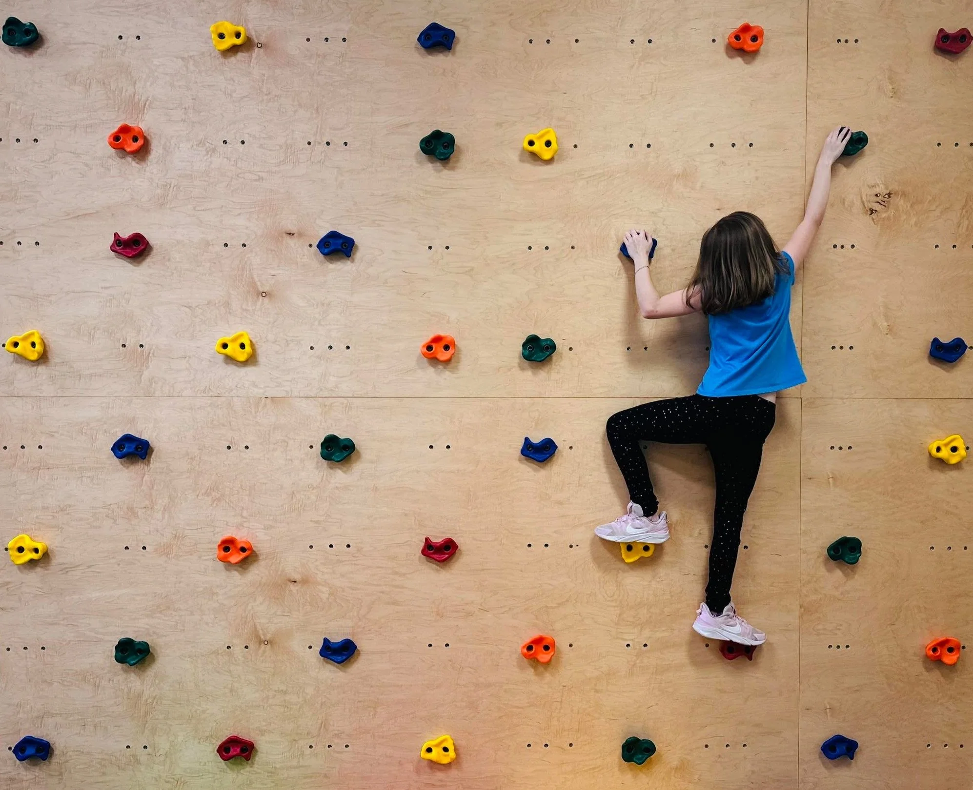 A young girl wearing a blue shirt, black pants with white polka dots, and pink shoes climbing a wooden indoor rock wall with colorful holds.