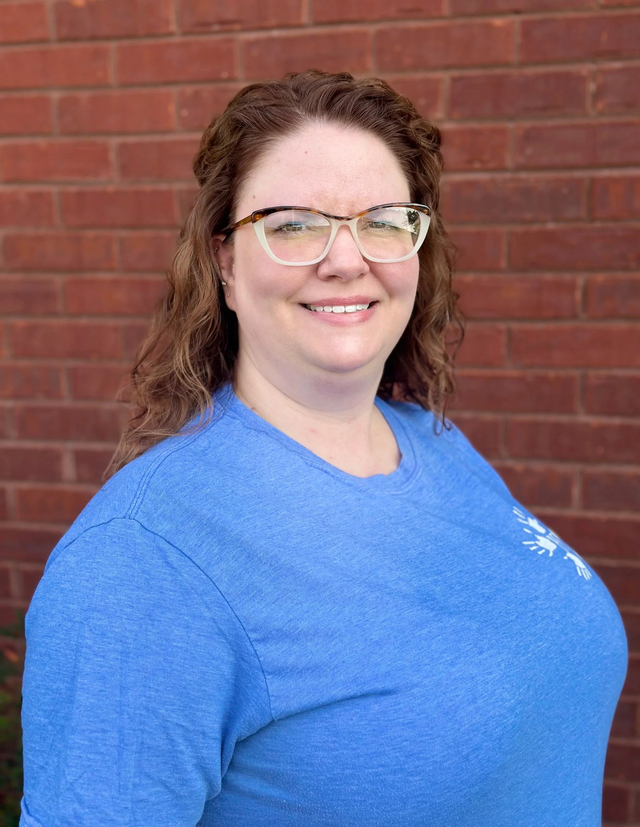 A woman with curly red hair and glasses smiling, wearing a blue shirt, standing in front of a red brick wall.