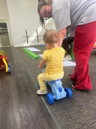 Child riding a toy vehicle while an adult helps in a room with toys and a green balance beam.