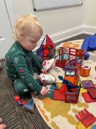 A young child playing with building blocks and toys on a yellow and orange rug in a playroom.