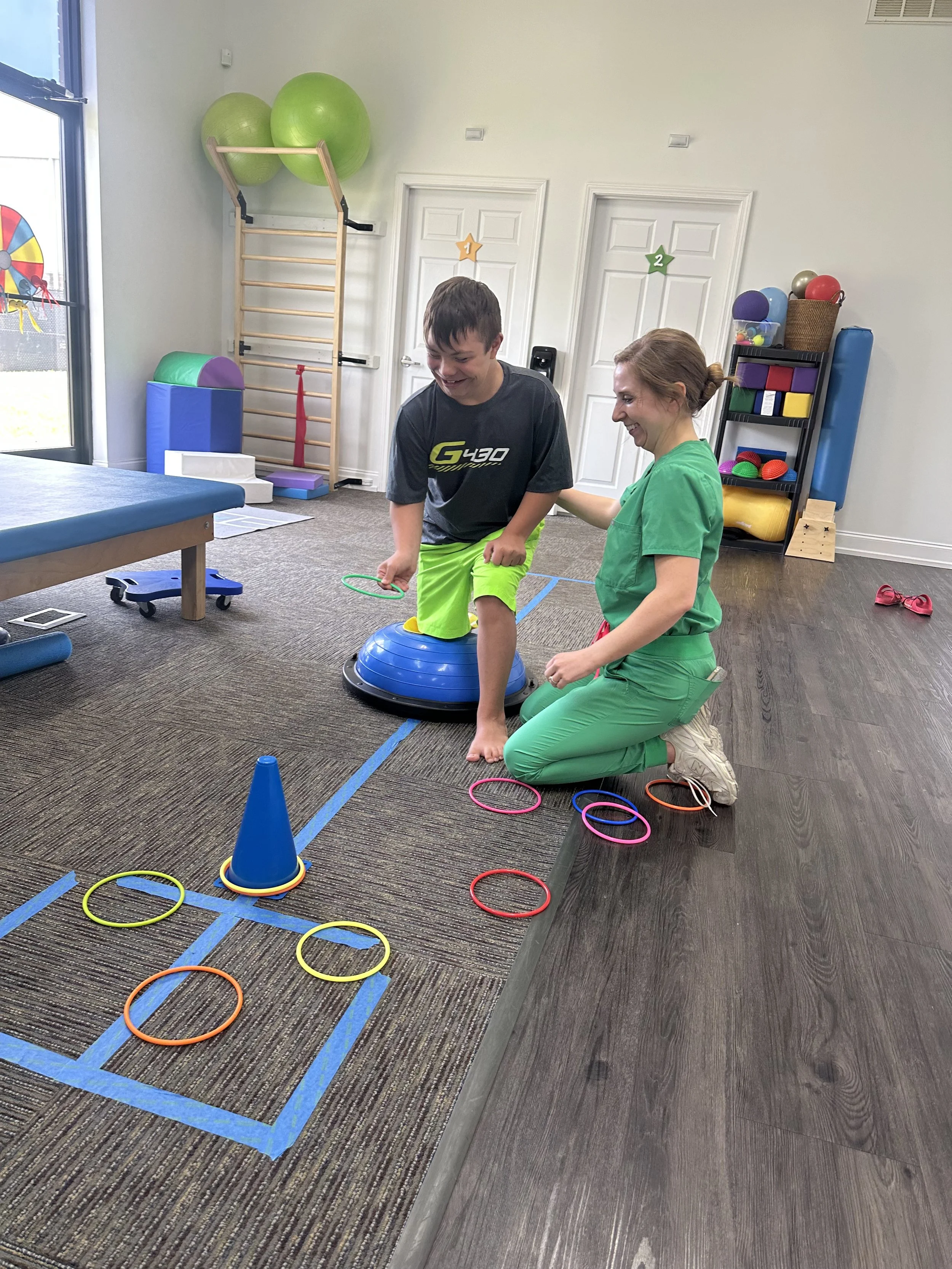 A young boy and a woman engage in an occupational therapy activity in a therapy room, with the boy balancing on a blue balance dome and the woman assisting him. The room has therapy equipment including exercise balls, foam blocks, and a storage shelf, with colorful hoops and a cone on the floor.