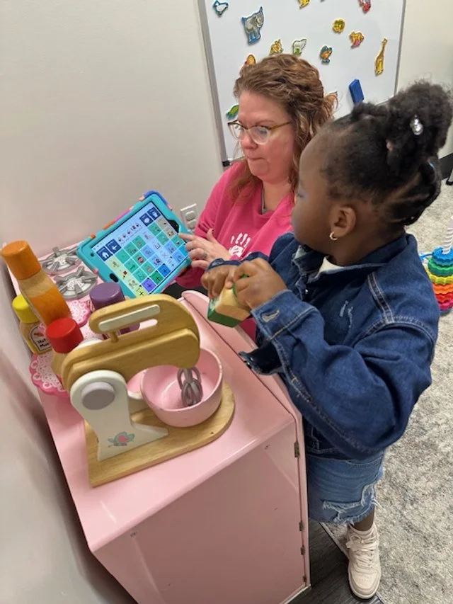 A woman and a young girl playing with a tablet at a pretend play kitchen set. The kitchen set has toy bottles and utensils on a pink surface.