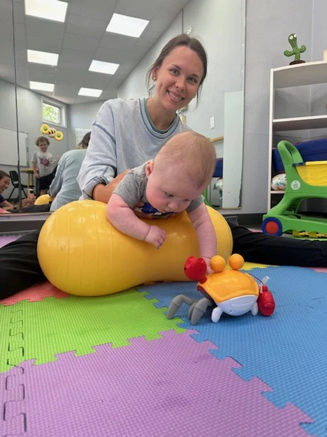 A woman and a baby playing on soft colorful foam mats in a therapy or playroom. The woman is smiling and the baby is on a yellow therapy ball with a toy crab nearby.