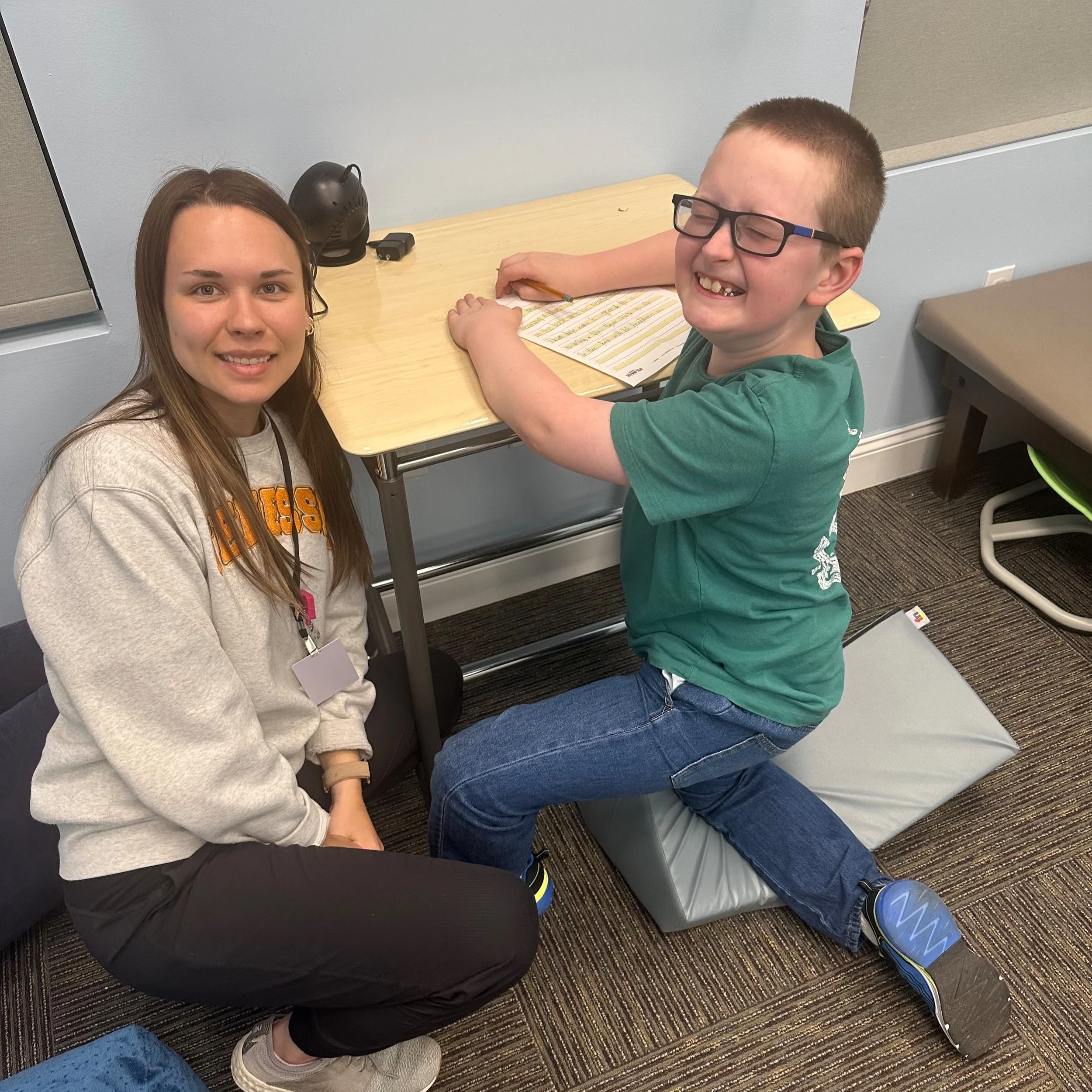 A girl and a boy are sitting together in a classroom; the girl is smiling at the camera and the boy, wearing glasses, appears to be laughing while writing on a piece of paper at a small table.