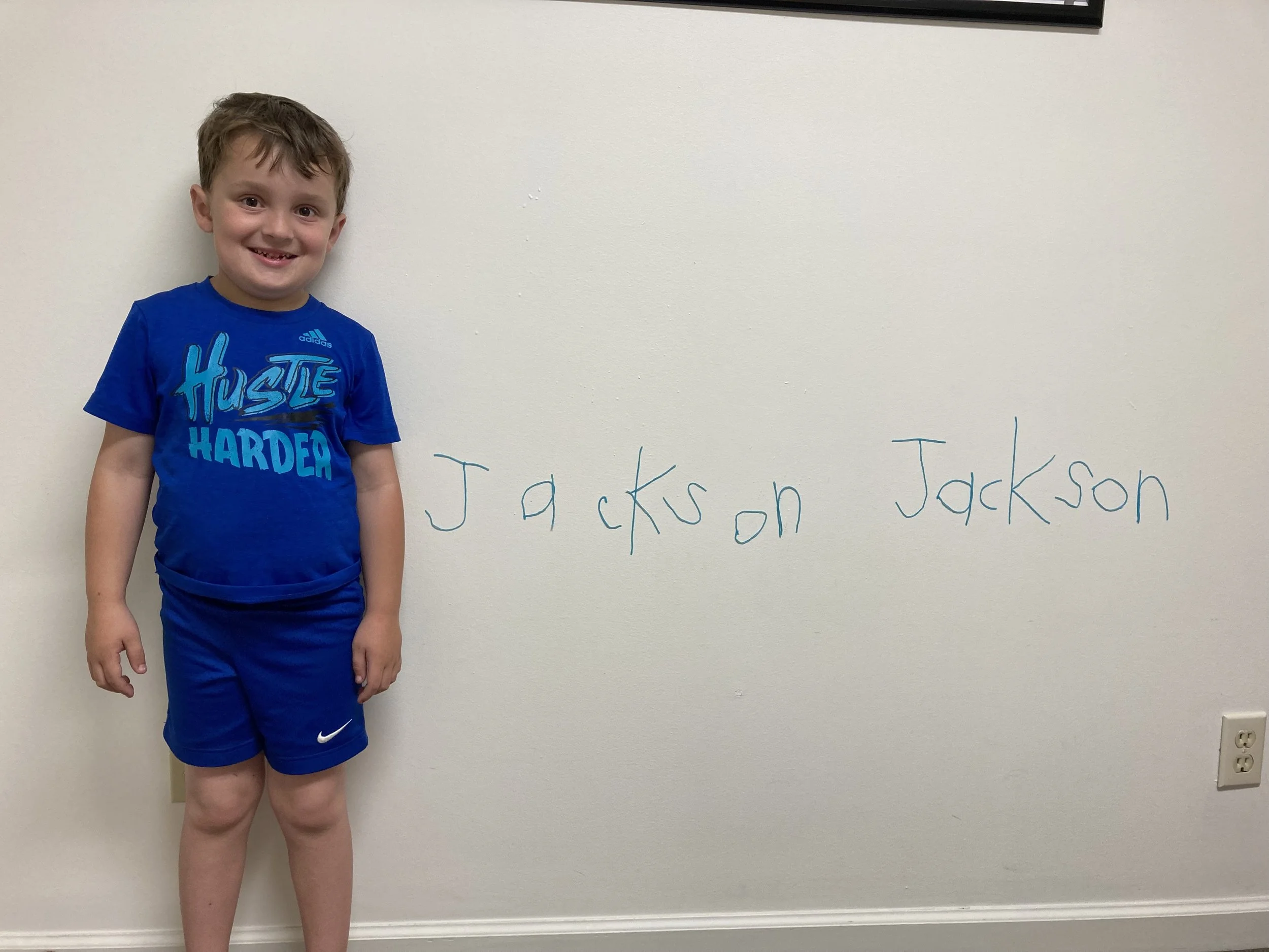A young boy wearing blue Nike and Adidas athletic clothing standing next to a white wall with the name 'Jackson Jackson' written on it in blue marker.