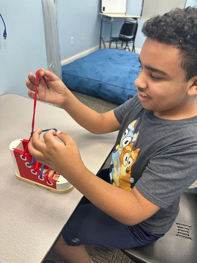 A young boy with curly hair playing a Connect 4 game at a table in a room with light blue walls and a bed in the background.