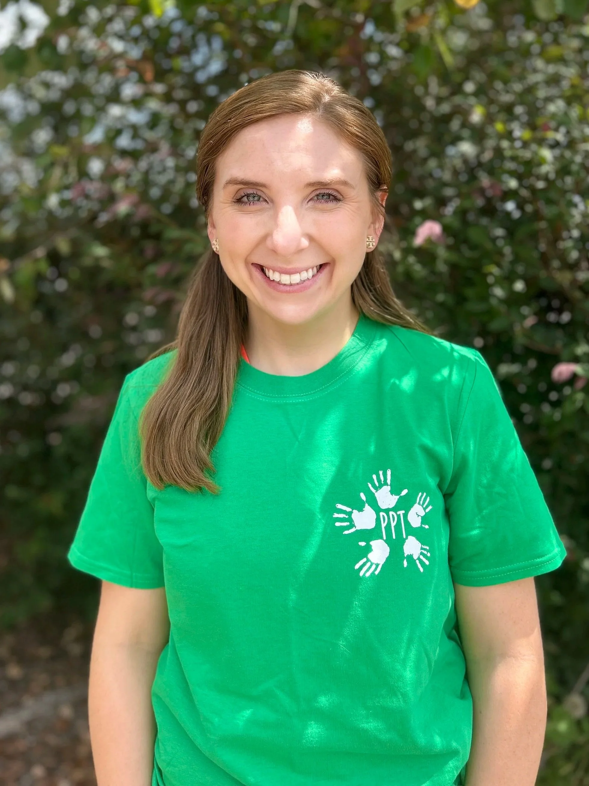 A smiling woman with light skin, long brown hair, and earrings, wearing a green t-shirt with a white logo of four handprints and the letters 'PPT', standing outdoors in front of a bush with green leaves and pink flowers.
