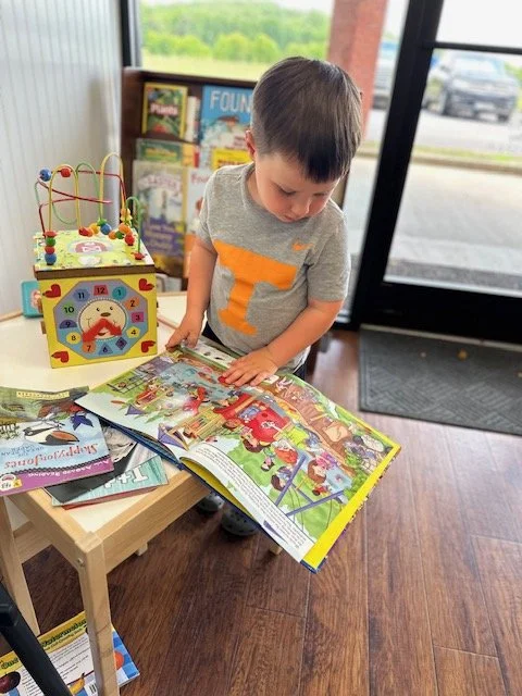 A young boy reading a colorful children's book at a small table inside a bookstore or library, near a window with a door outside visible.