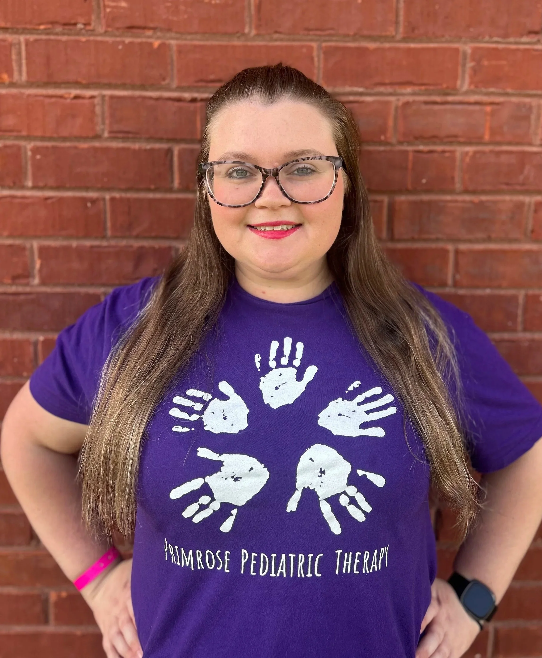Woman with long brown hair, glasses, and a purple shirt standing against a brick wall.