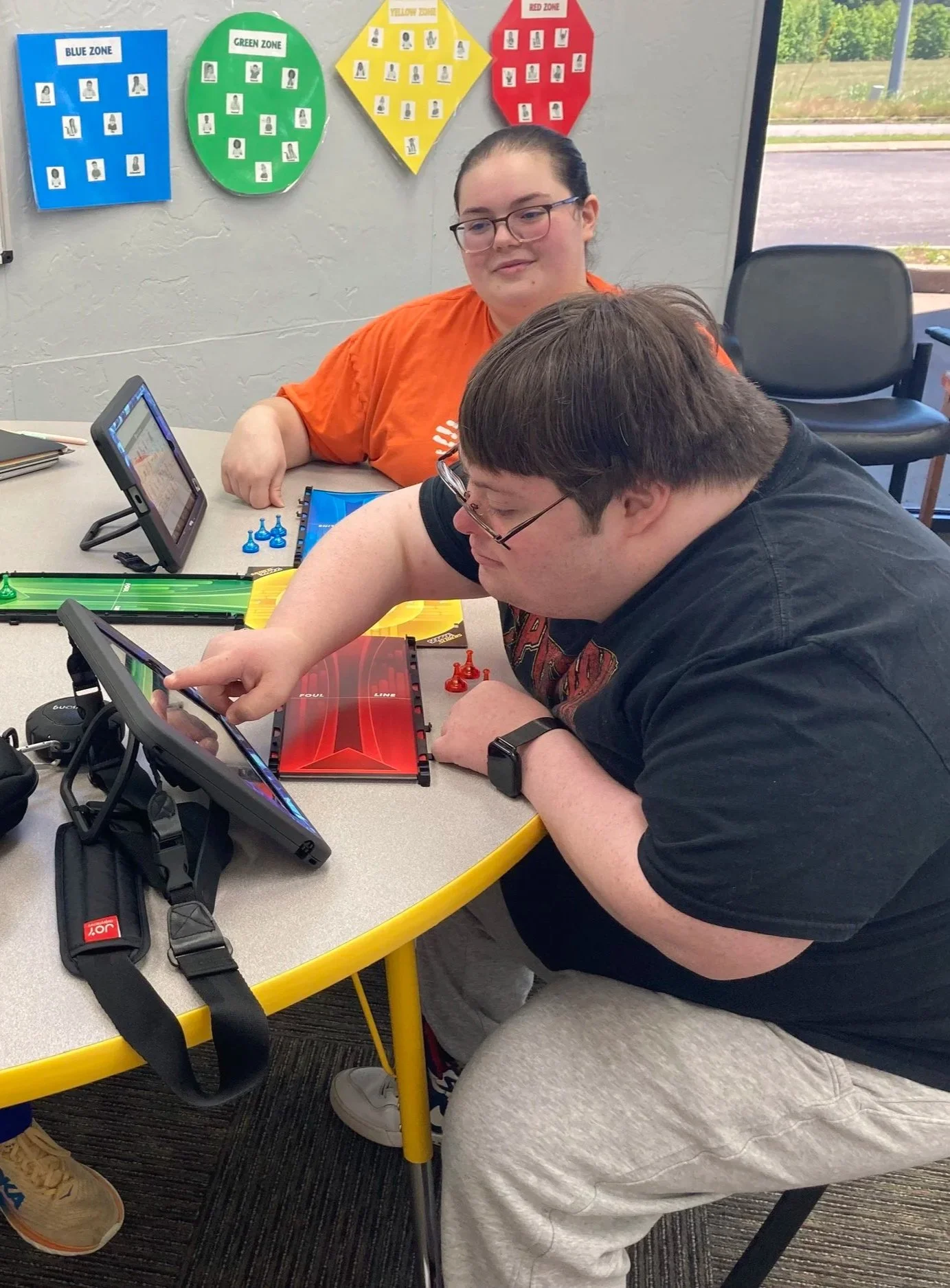 Two people playing a digital board game at a table in a classroom, with colorful zone posters on the wall behind them and a window to the outside.