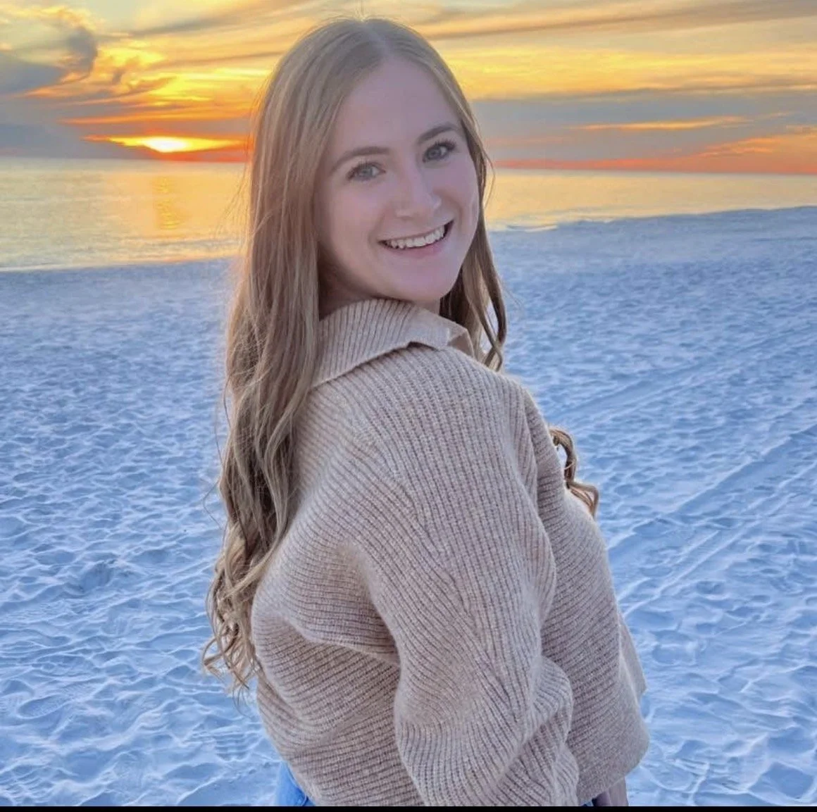 A young woman with long wavy hair smiling at the camera on a beach during sunset, wearing a beige sweater.