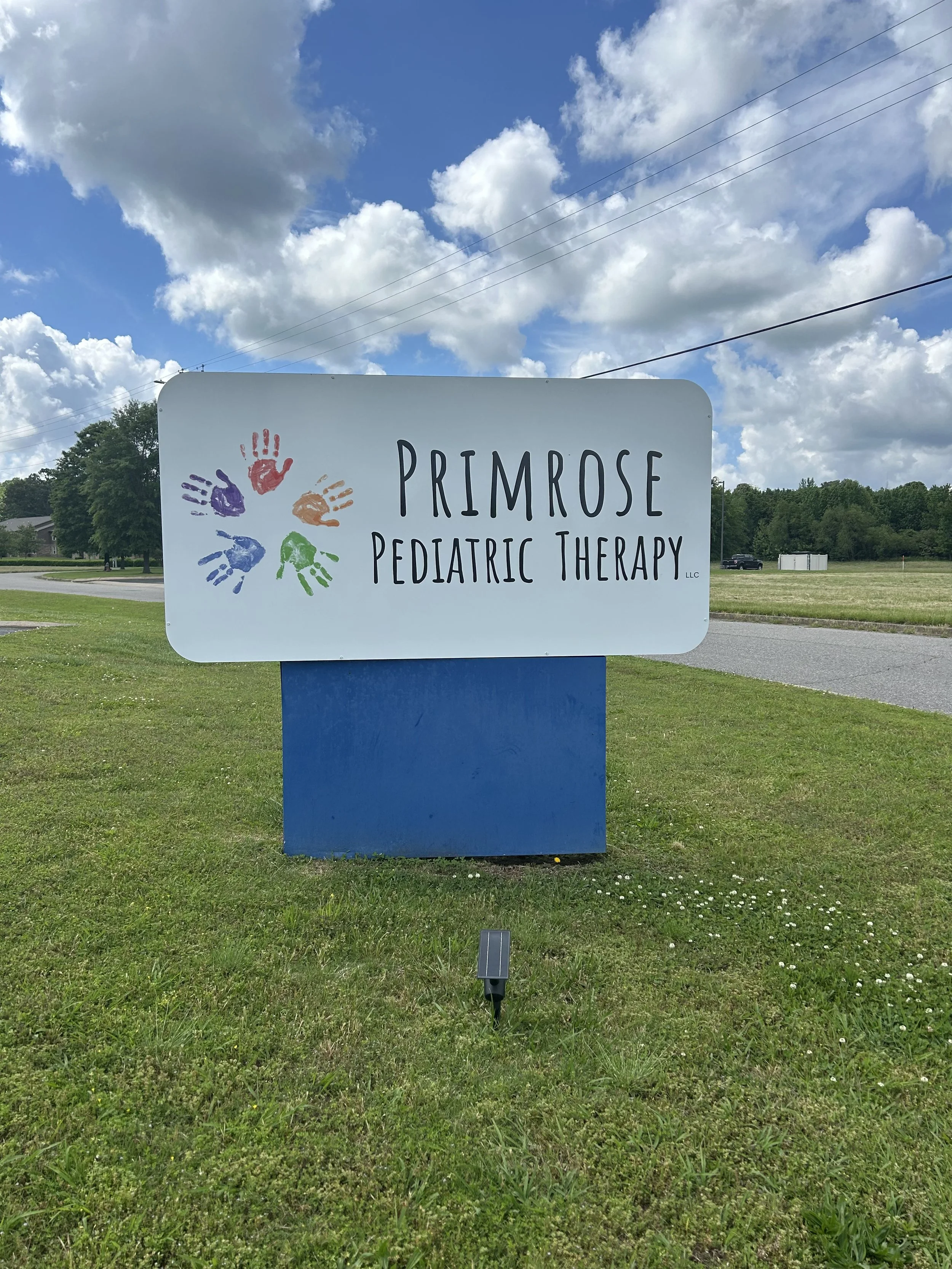 Sign for Primrose Pediatric Therapy with colored handprints on the left side and a partly cloudy sky in the background.