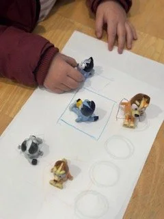 Child's hand drawing on paper with small animal figurines including a black and white dog, a gray dog, a brown and white dog, and a yellow and brown dog.