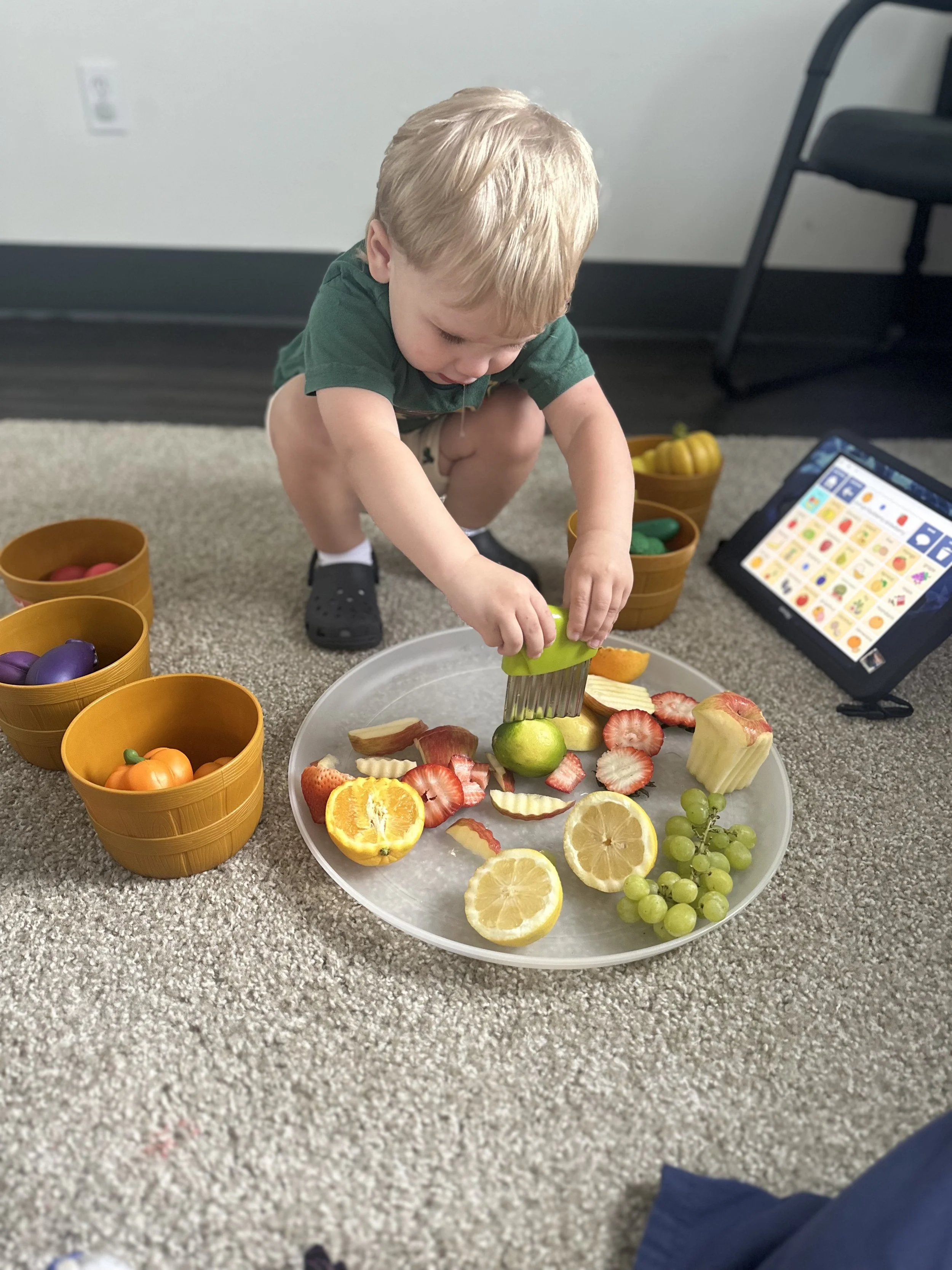 A young boy playing with toy food on a tray, pretending to cut fruit with a toy knife, surrounded by bowls of colorful plastic fruits, with a tablet displaying educational content nearby.