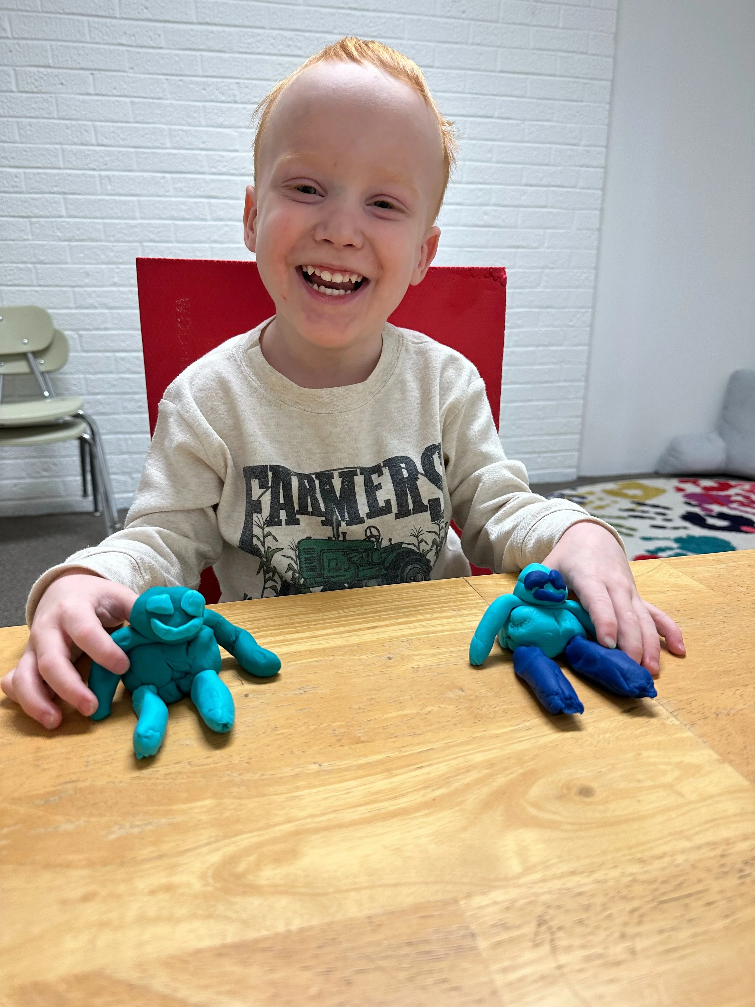 A young boy sitting at a wooden table with a big smile, holding two clay sculptures of humanoid creatures, one in each hand. The sculptures are made of blue and teal clay and have facial features and limbs.
