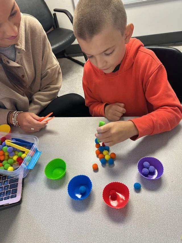 Two children playing with colorful pom-poms and silicone cupcake liners on a table in a classroom setting.