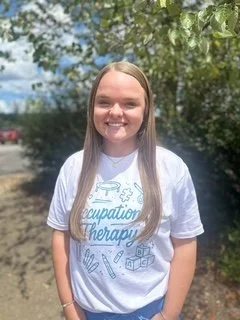 Young woman with long blonde hair, smiling outdoors in a park, wearing a white T-shirt with blue text and graphics.