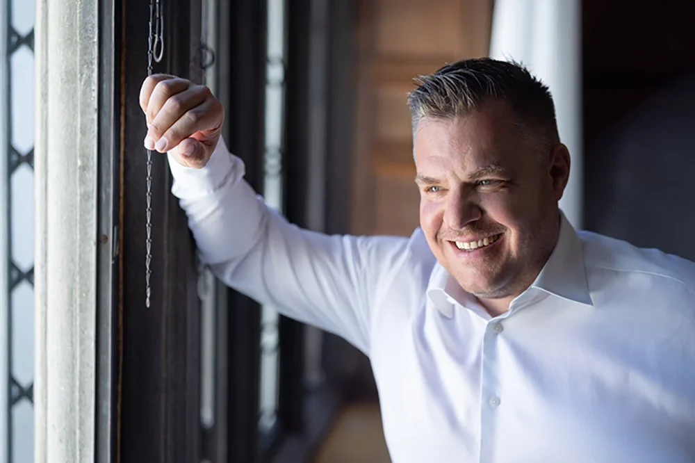 Lächelnder Mann in weißem Hemd lehnt an einem Fenster mit Lamelle, bei Tageslicht.