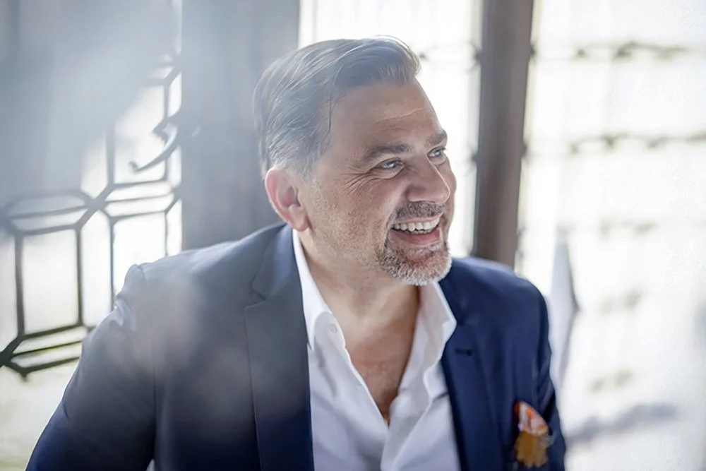 Ein lächelnder Mann in einem blauen Anzug und weißem Hemd sitzt an einem Tisch in einem hell erleuchteten Raum.
