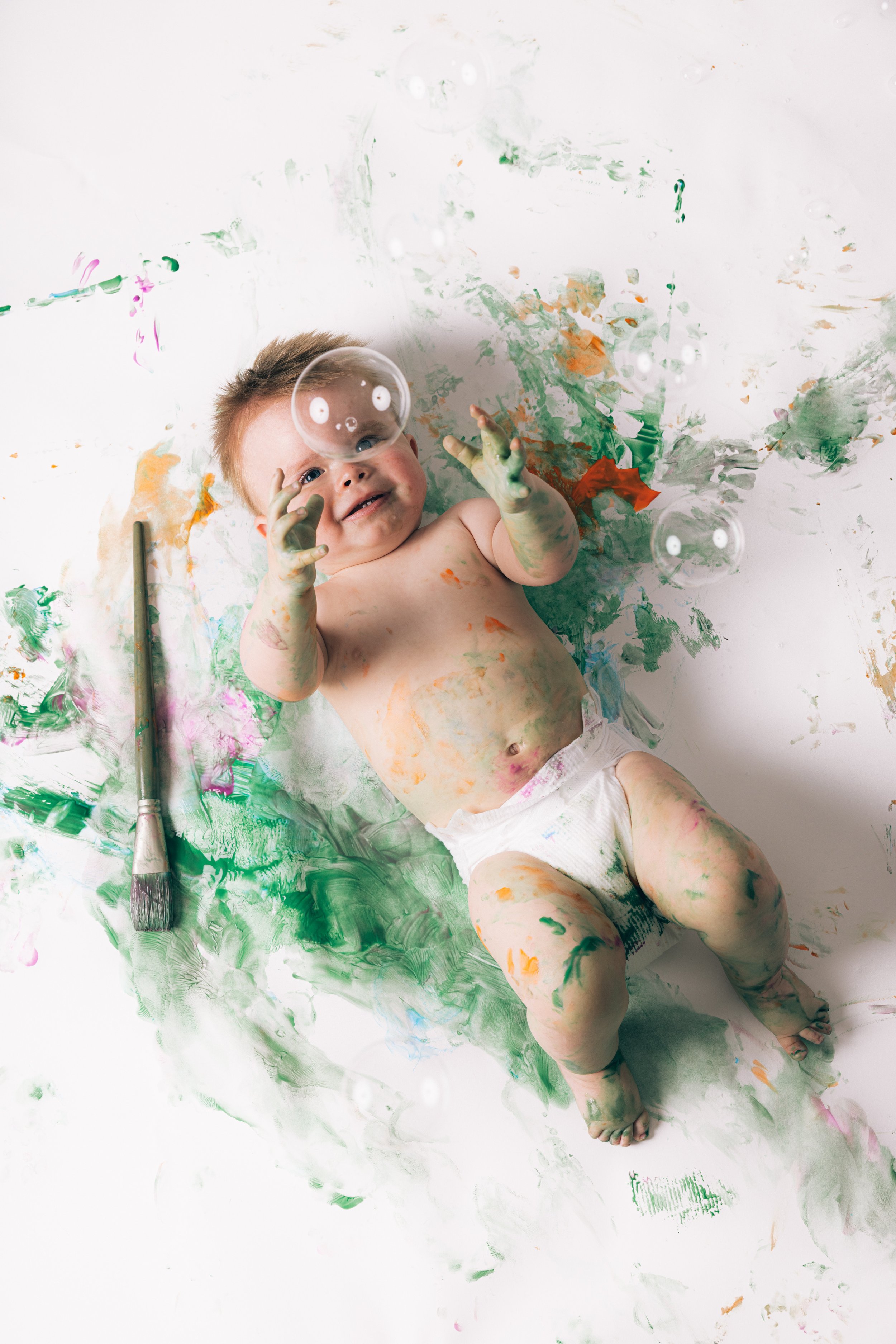 A young child with messy hair lying on a white surface covered in multicolored paint, reaching for a transparent bubble, with a paintbrush nearby.