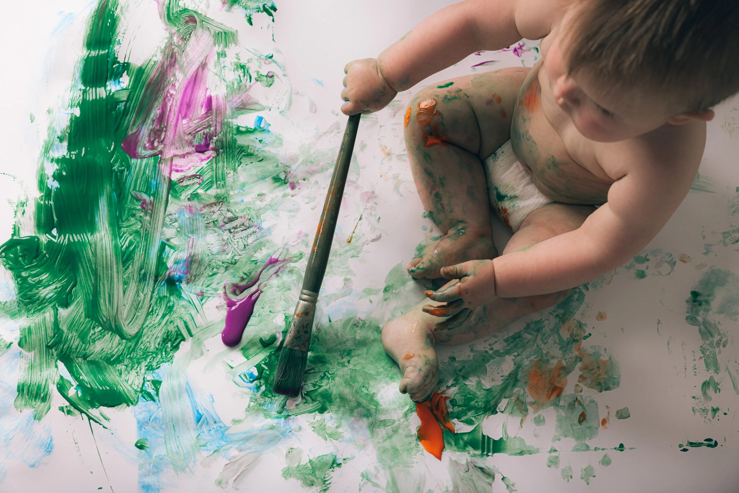 Two young children with paint on their bodies and hands, sitting on a white surface, playing with colorful paint, using their hands and a paintbrush.