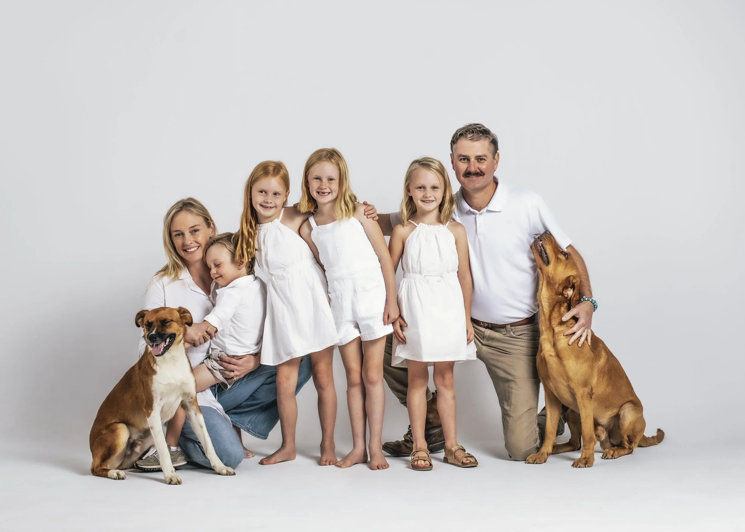 A family of six, including four young girls, a woman, a man, and two dogs, pose together against a plain white backdrop. They are all smiling and dressed in white clothing, with the dogs sitting and being petted.
