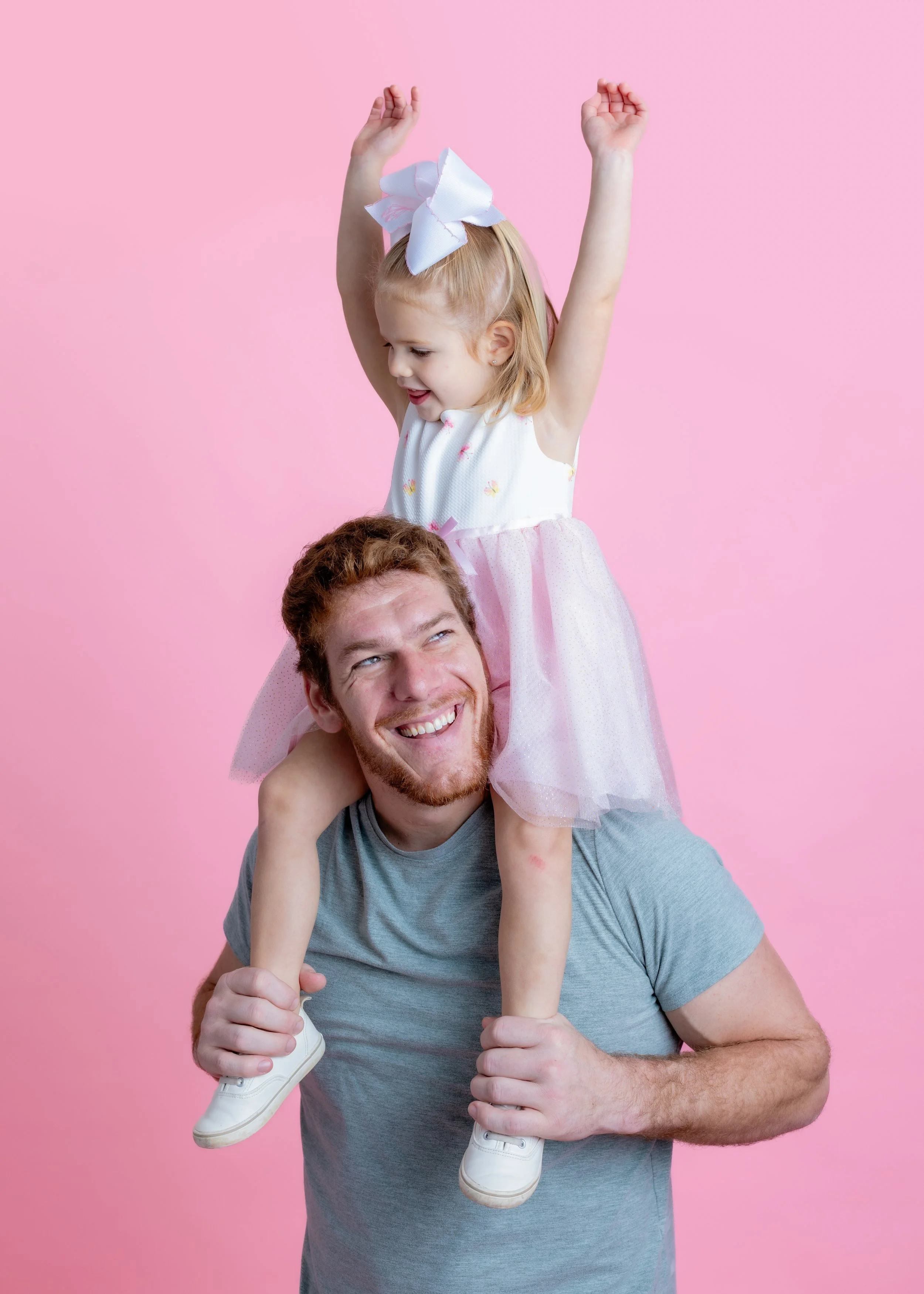 A man with a beard and short hair smiling while carrying a young girl on his shoulders. The girl has blonde hair with a large white bow, wearing a white and pink dress, raising her arms joyfully against a pink background.