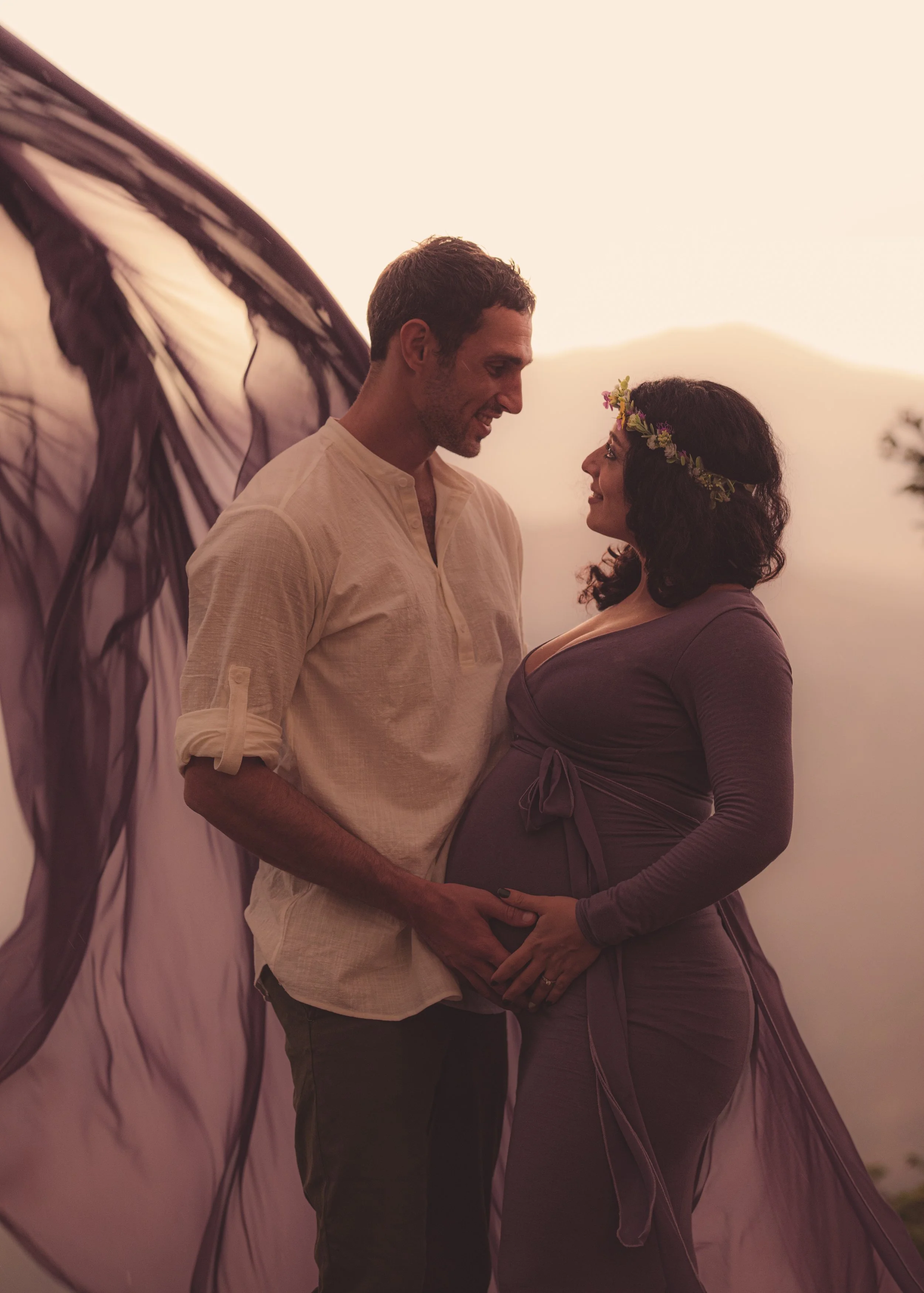 A couple standing close together, touching their baby bump, with the man gently holding the woman's belly, during sunset or sunrise.
