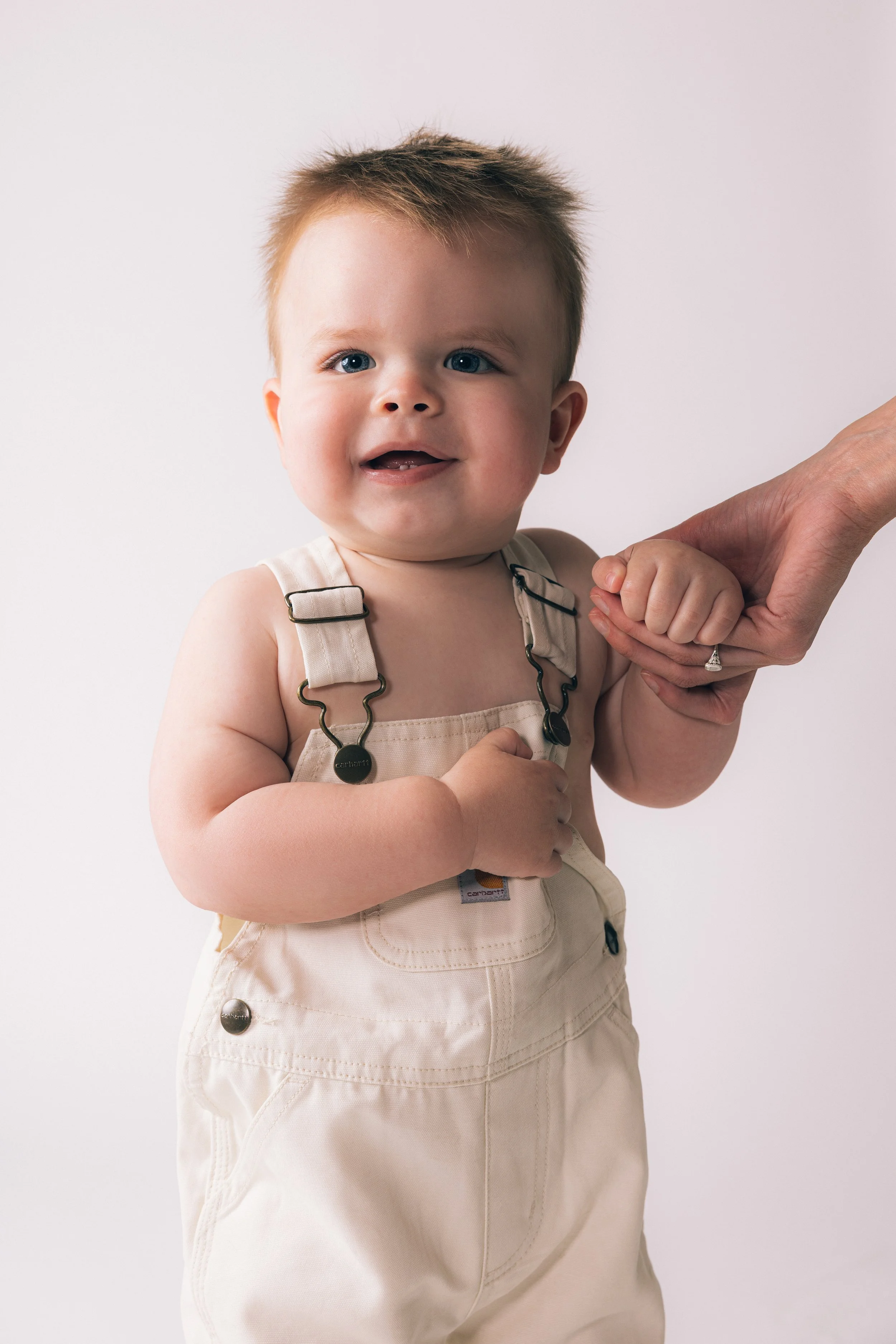 A smiling toddler with blue eyes and light brown hair holds hands with an adult, possibly the child's parent, against a plain white background. The child is wearing beige overalls with metal clasps.