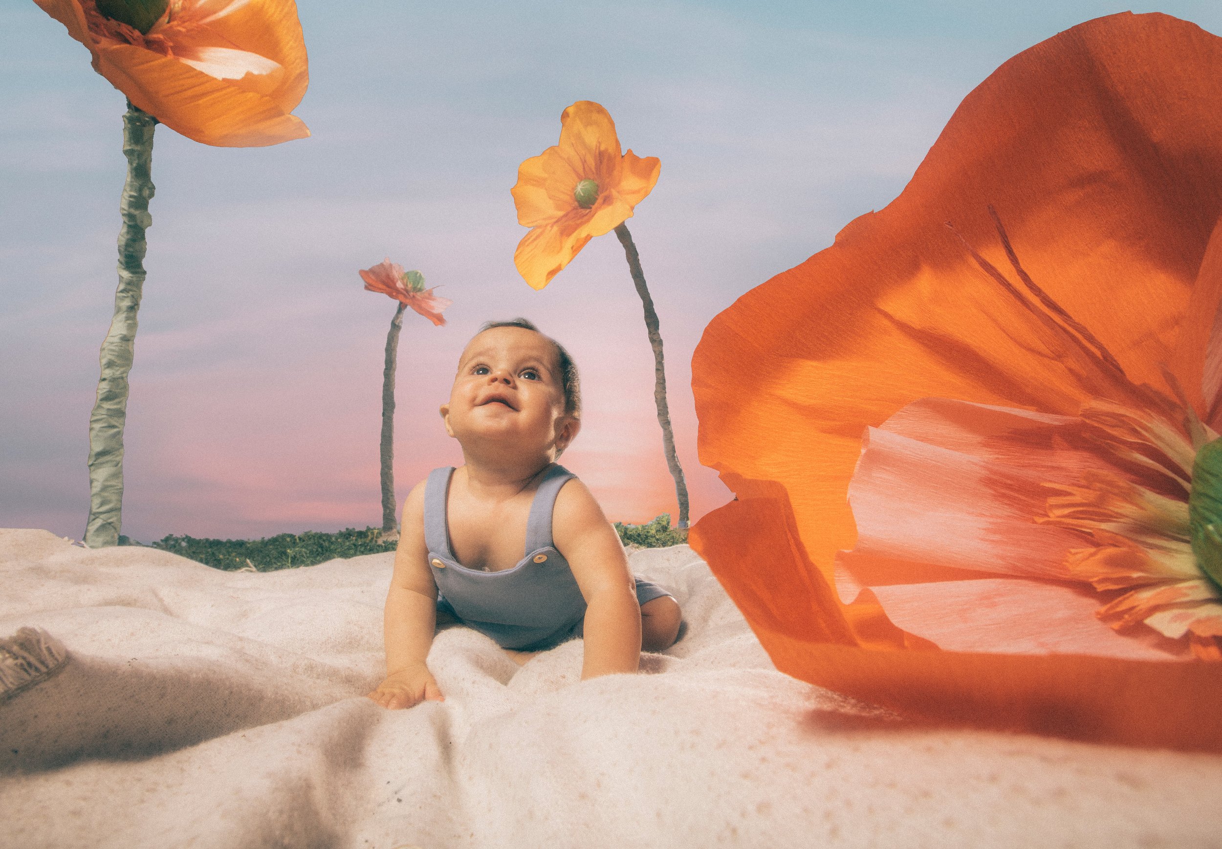 A baby crawling on a sandy surface with large orange flowers in the foreground and a pastel-colored sky with trees in the background.