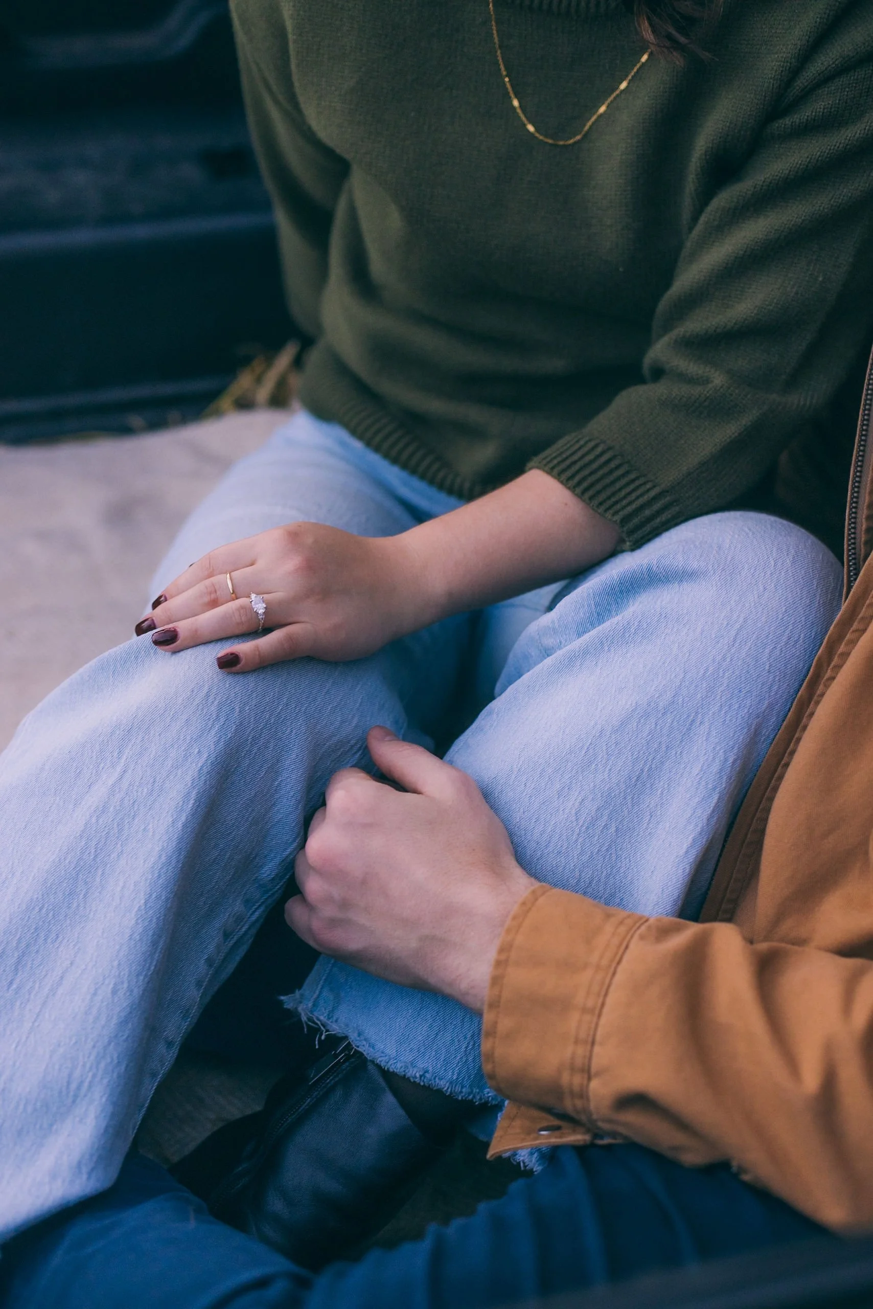 A person wearing a green sweater and jeans has their hand resting on their lap, showing an engagement ring. Someone else, in a brown jacket, has their hand gently placed on the person's other knee, creating an intimate moment.