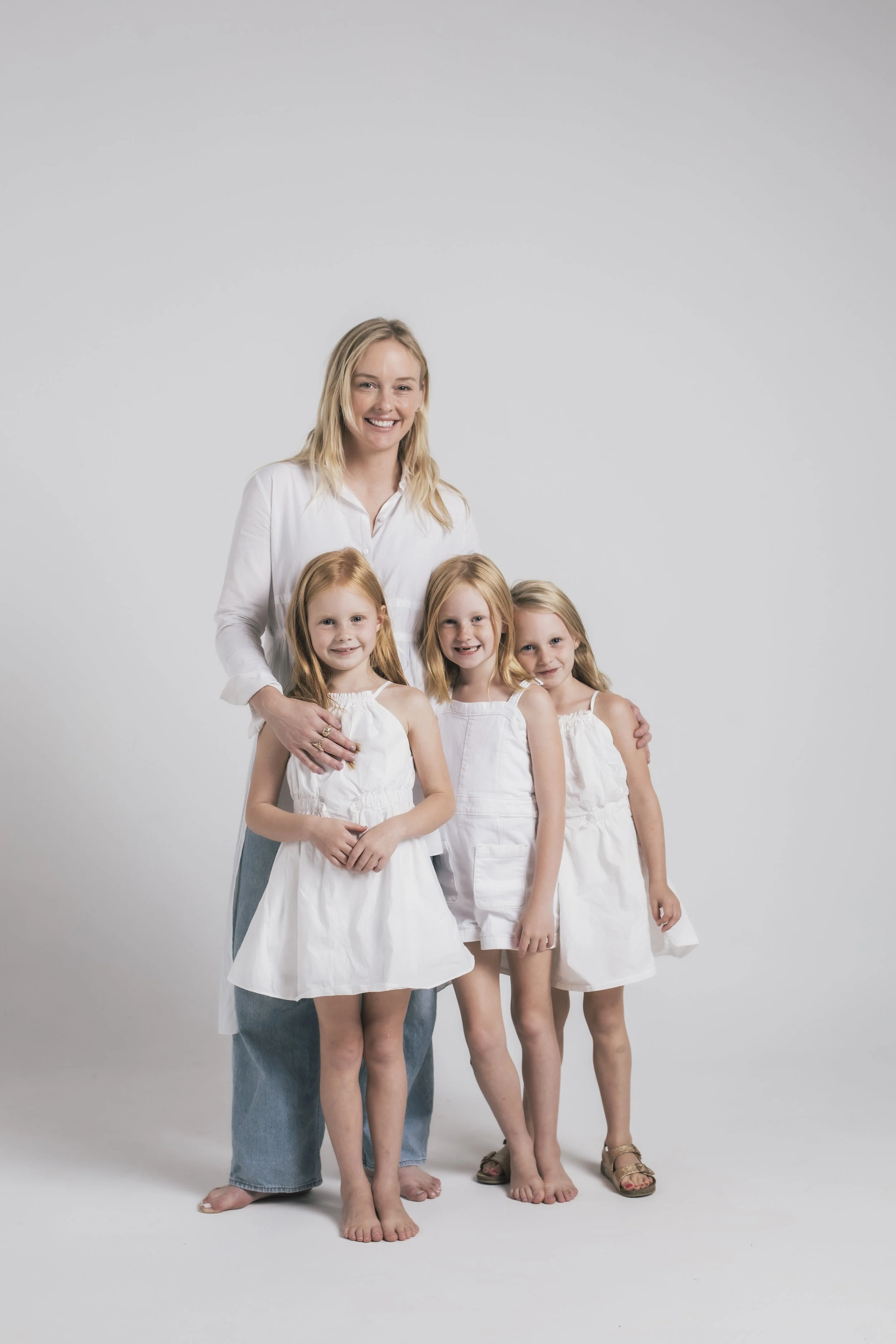 A smiling woman standing with three young girls, all dressed in white, against a plain white background.