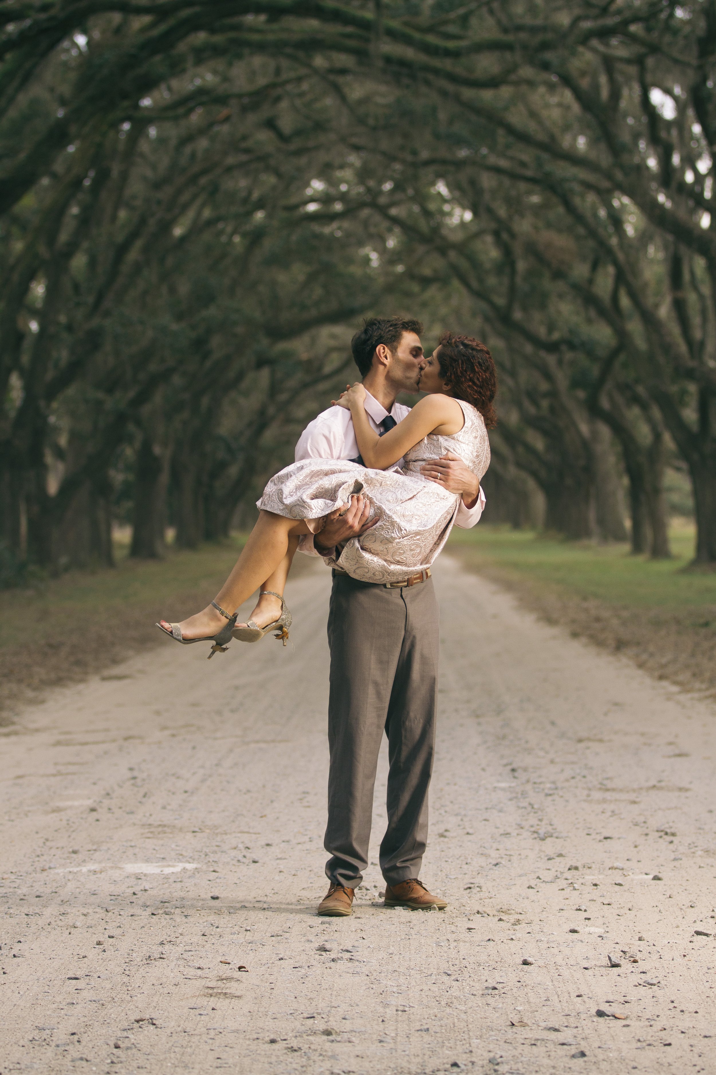 A man in dress pants and a shirt holding a woman in a pink dress, both kissing under an archway of trees on a dirt road.