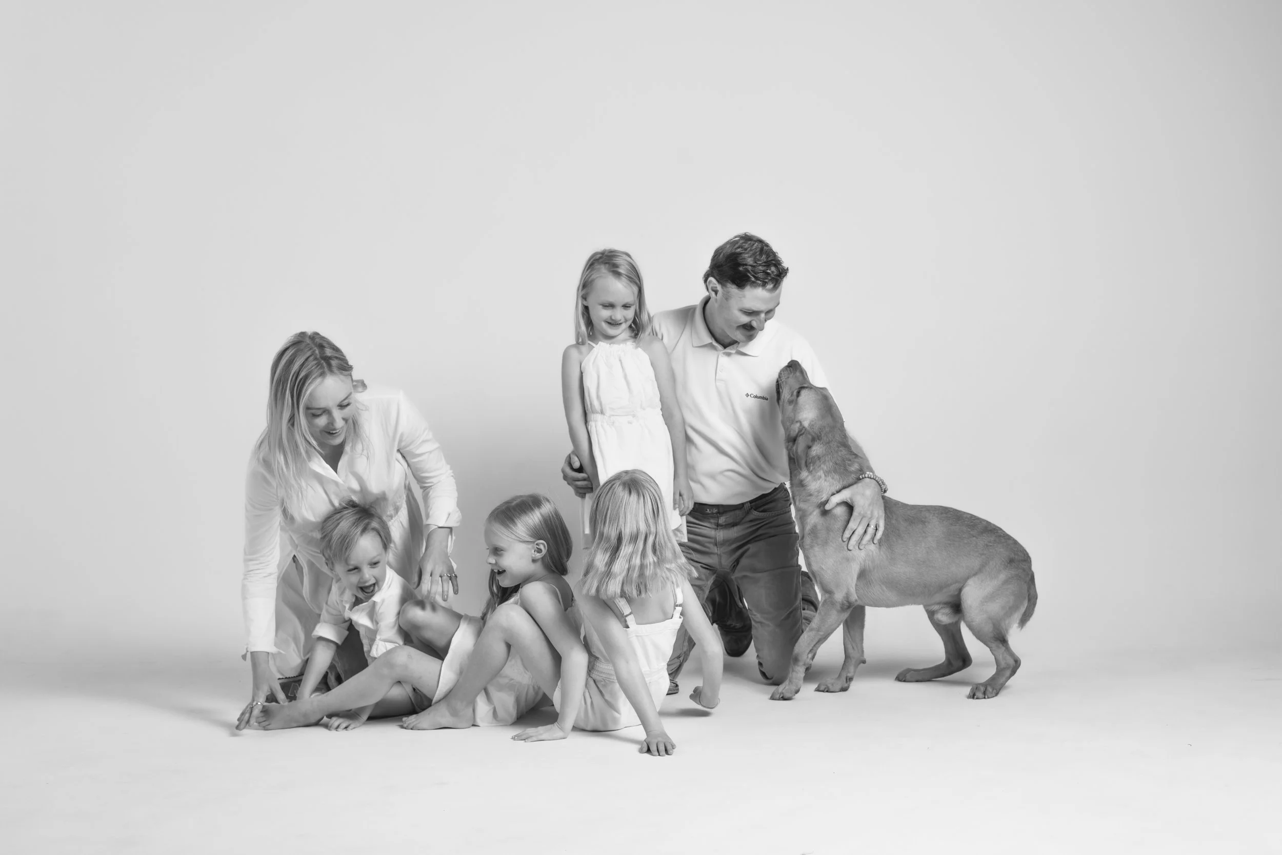 Family of six, including two adults and four children, with a large dog, playing and smiling in a studio with a plain background.
