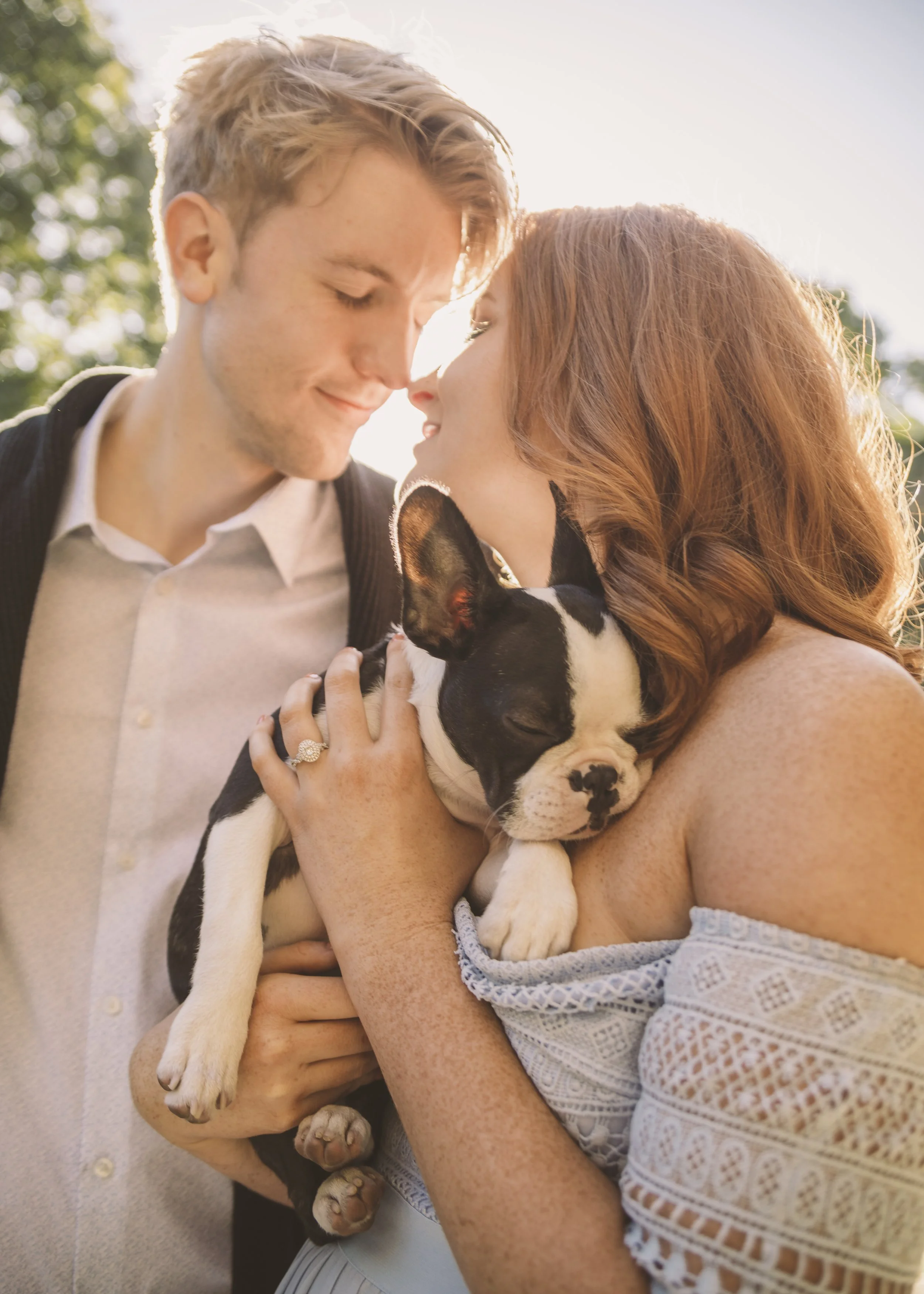 A couple is holding a sleeping Boston Terrier puppy close to their faces outdoors during sunset, with trees in the background.