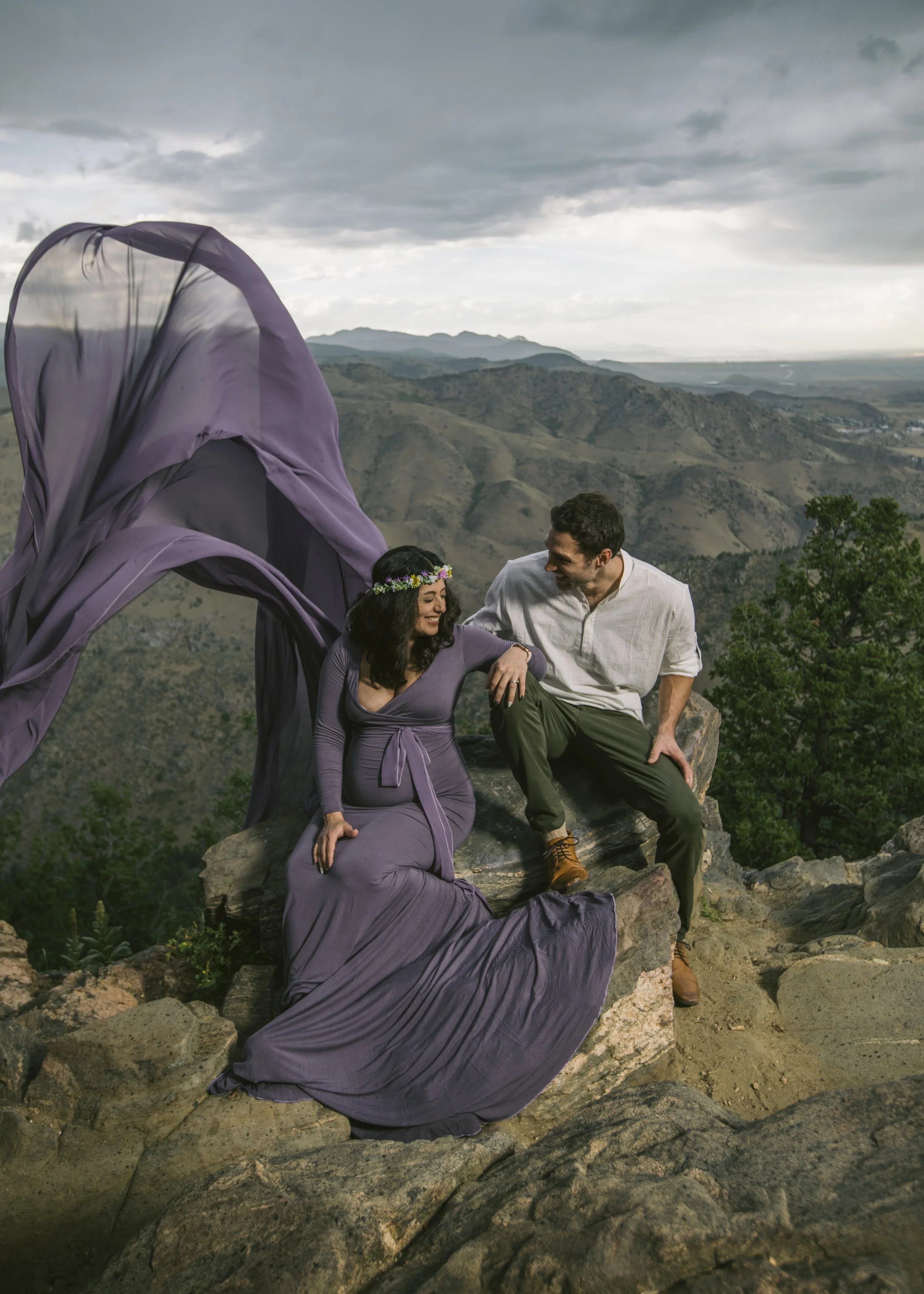 A couple sitting on rocks outdoors, smiling and holding hands with a scenic mountain landscape in the background. The woman wears a purple dress and a flower crown, and the man wears a white shirt and green pants.
