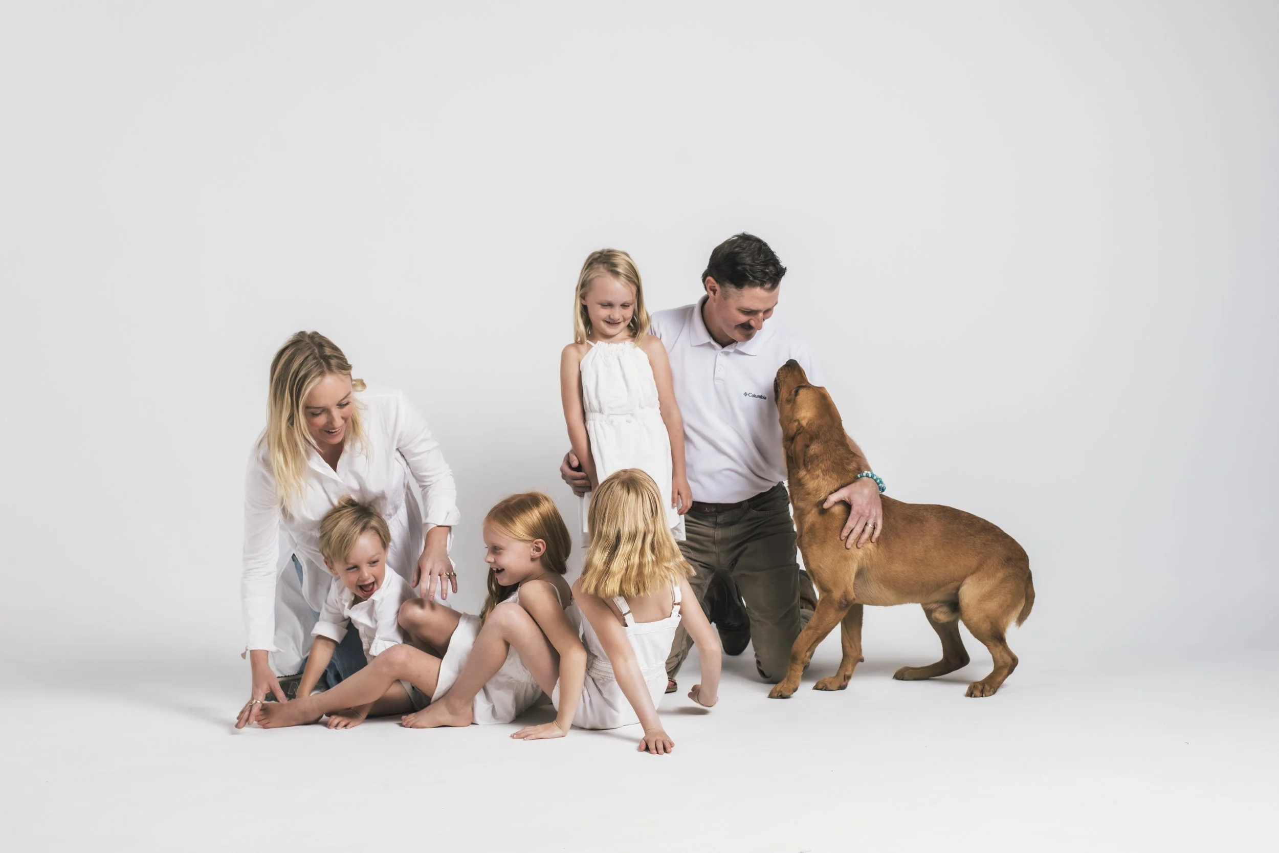 Family of five, including two women, two young girls, and a man, with a large brown dog, all smiling and interacting in a white studio setting.