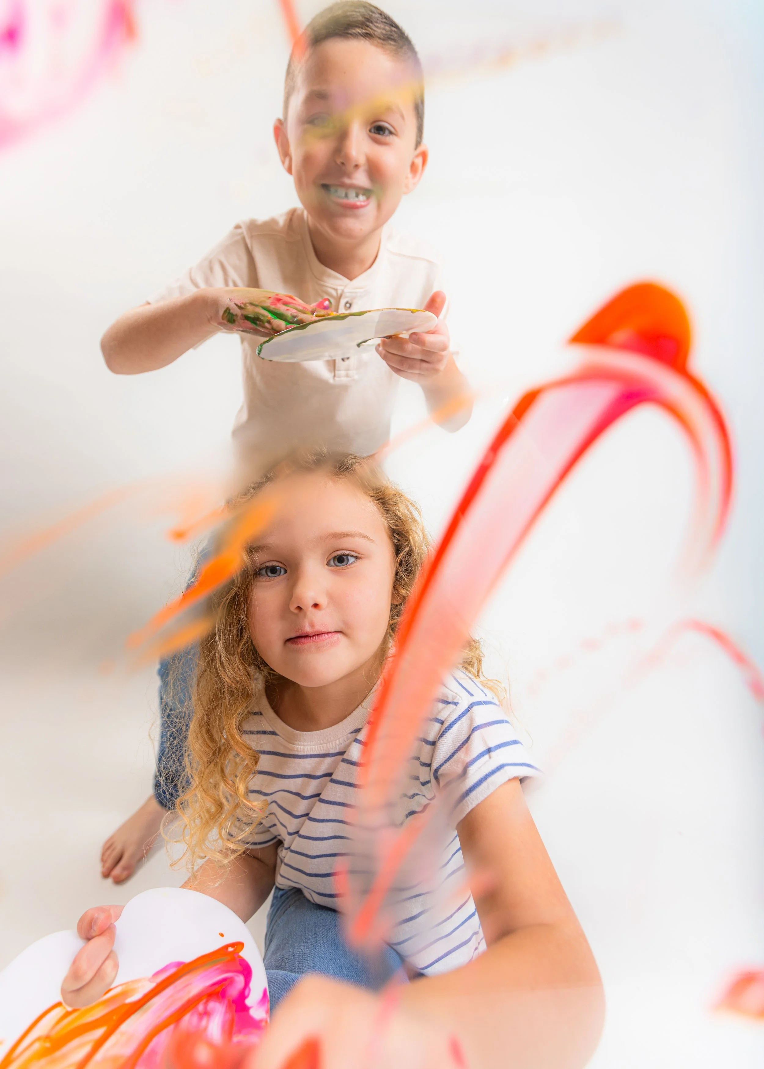 Two children, a boy and a girl, playing with colorful ribbons and paint on a transparent surface. The boy is standing, holding a plate with painted fingers, and smiling. The girl is sitting, looking at the camera with a neutral expression, and the ri