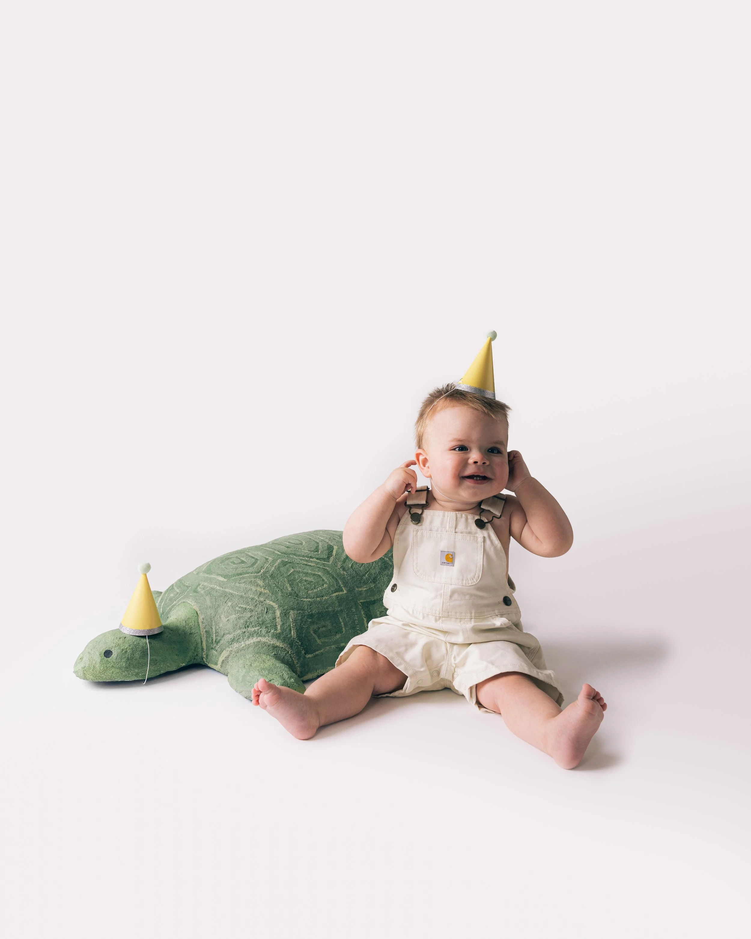 A young child sitting on the floor wearing a party hat and beige overalls with a crocodile plush toy beside them, also wearing a party hat, against a plain white background.