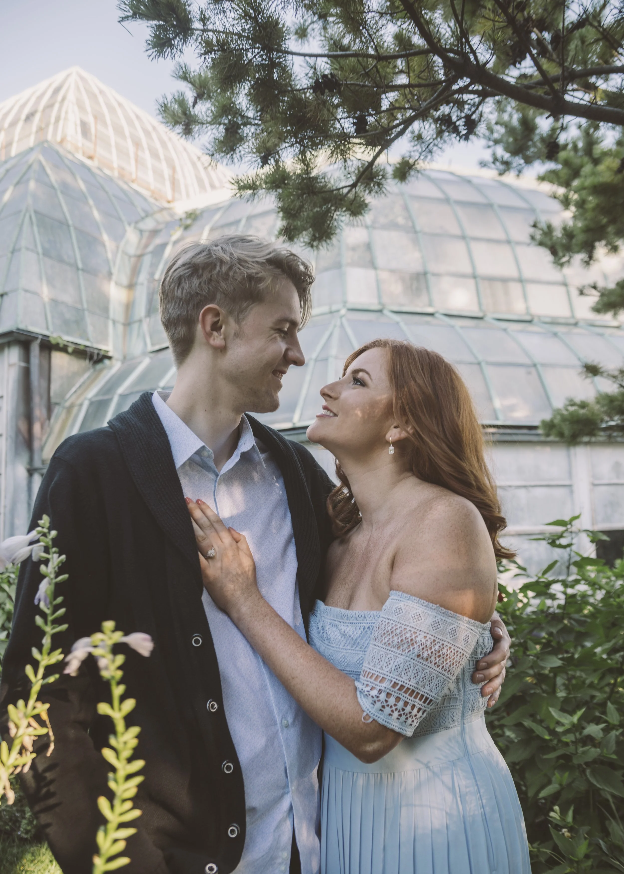 A couple gazes lovingly into each other's eyes amid greenery and a glass greenhouse structure in the background.