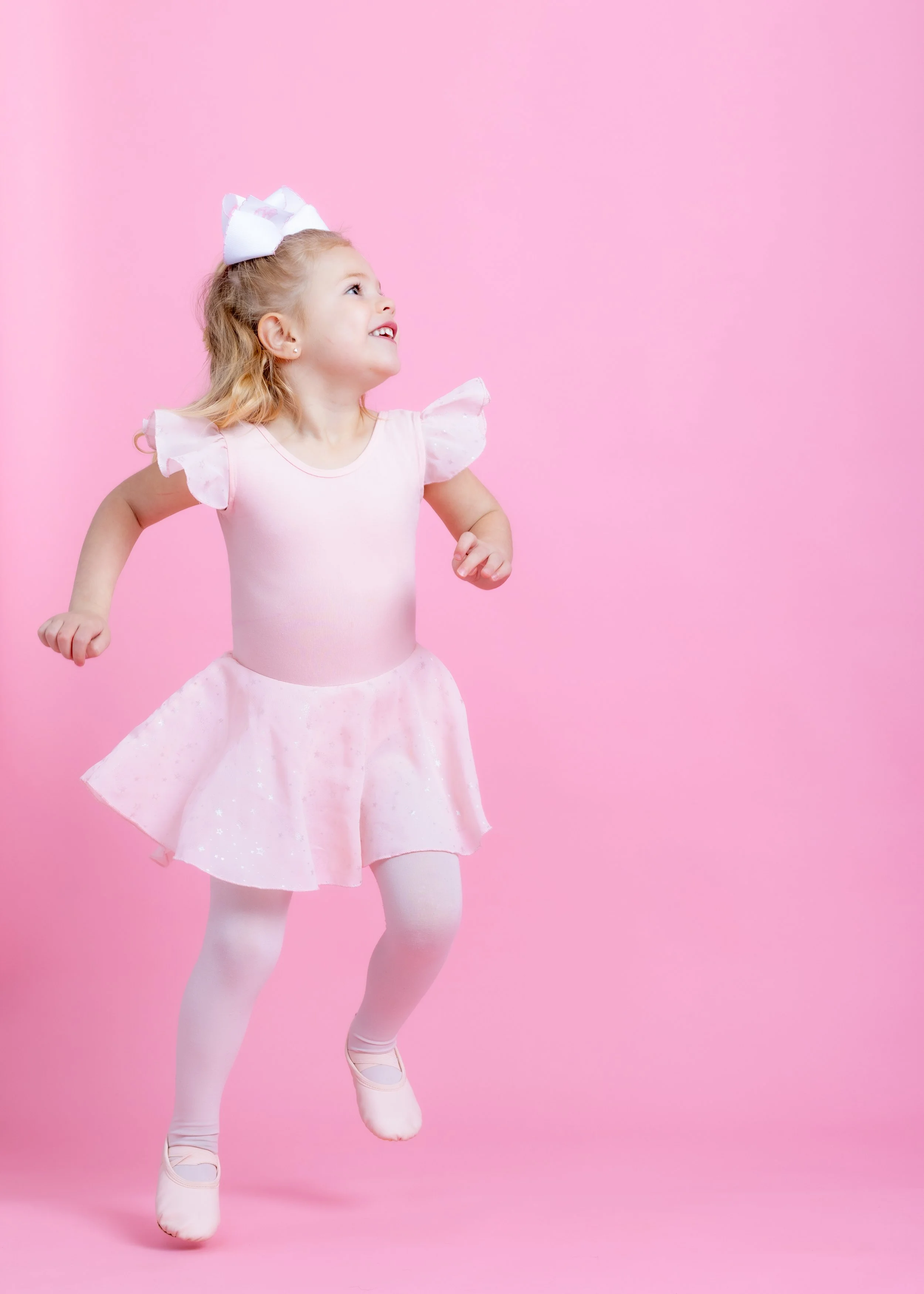 Young girl in a pink ballet outfit jumping against a pink background.
