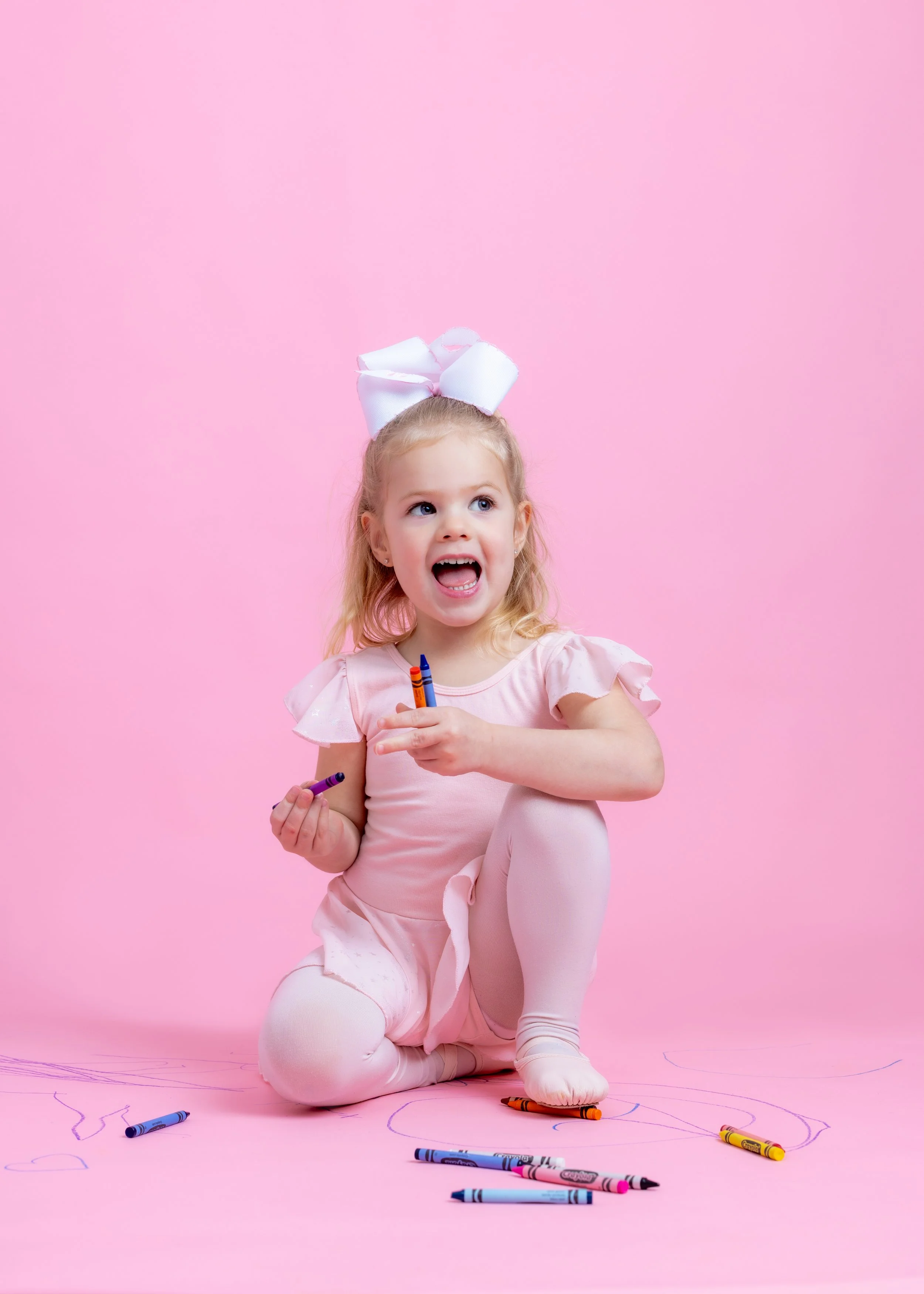 A young girl in pink dress and white tights sitting on a pink background, holding crayons and surrounded by crayon drawings.