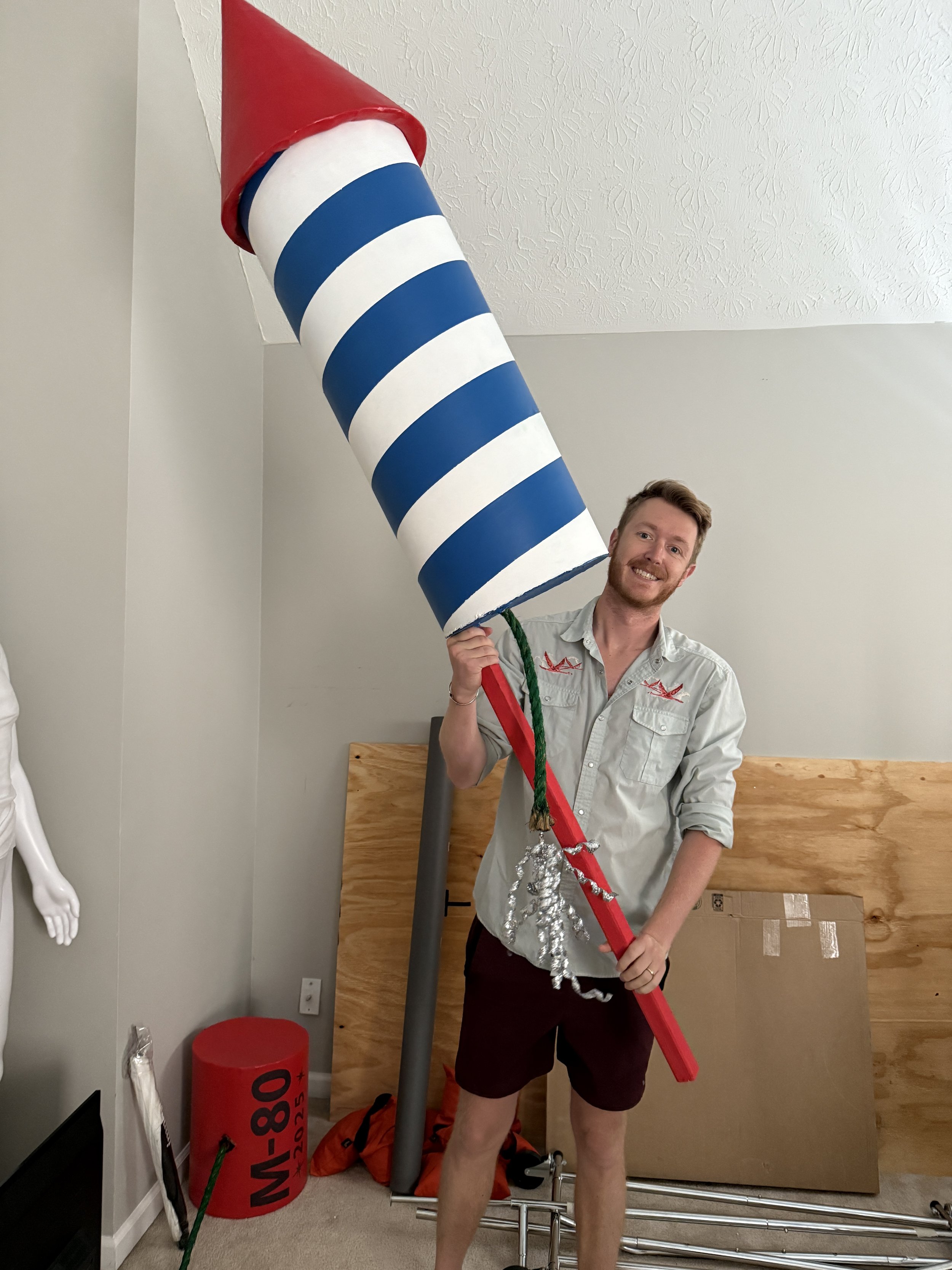A man smiling and holding a large, striped, cylindrical object resembling a candy cane or a rocket, indoors with various objects including a red cylindrical object with a label, a cardboard, and some metallic poles.
