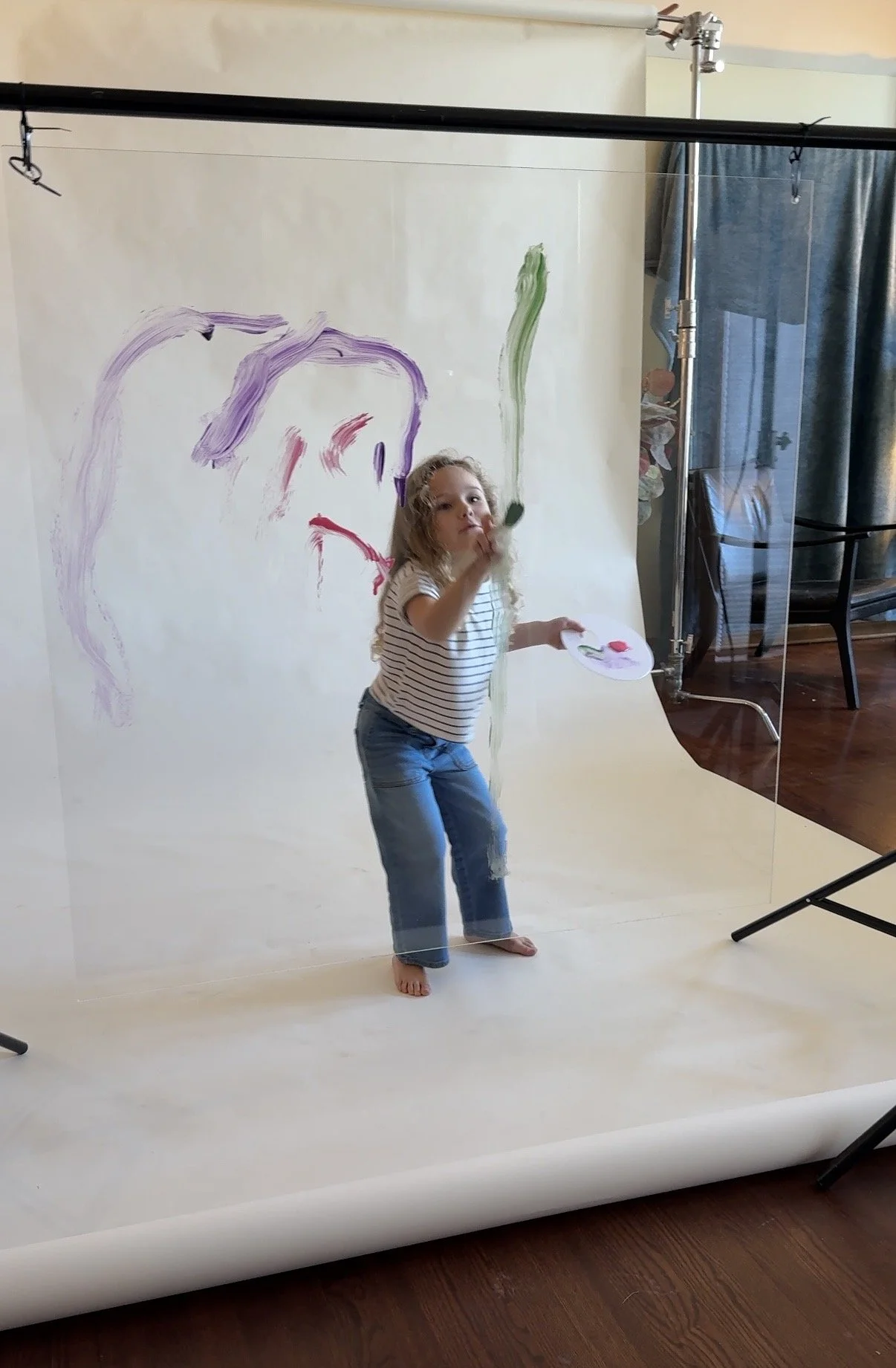 Young girl with long curly hair standing in front of a photography backdrop, painting a large face with colorful brushstrokes. She is holding a palette and applying paint.
