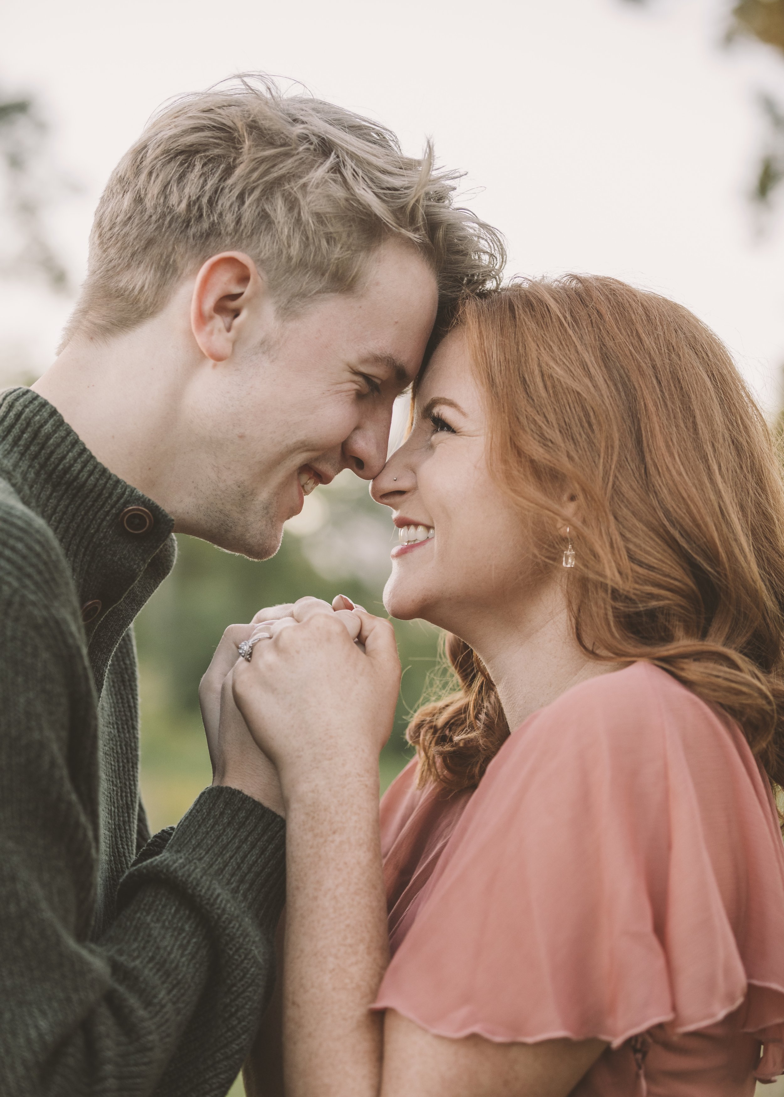 A smiling couple with their foreheads and noses touching, holding hands, outdoors during daytime.