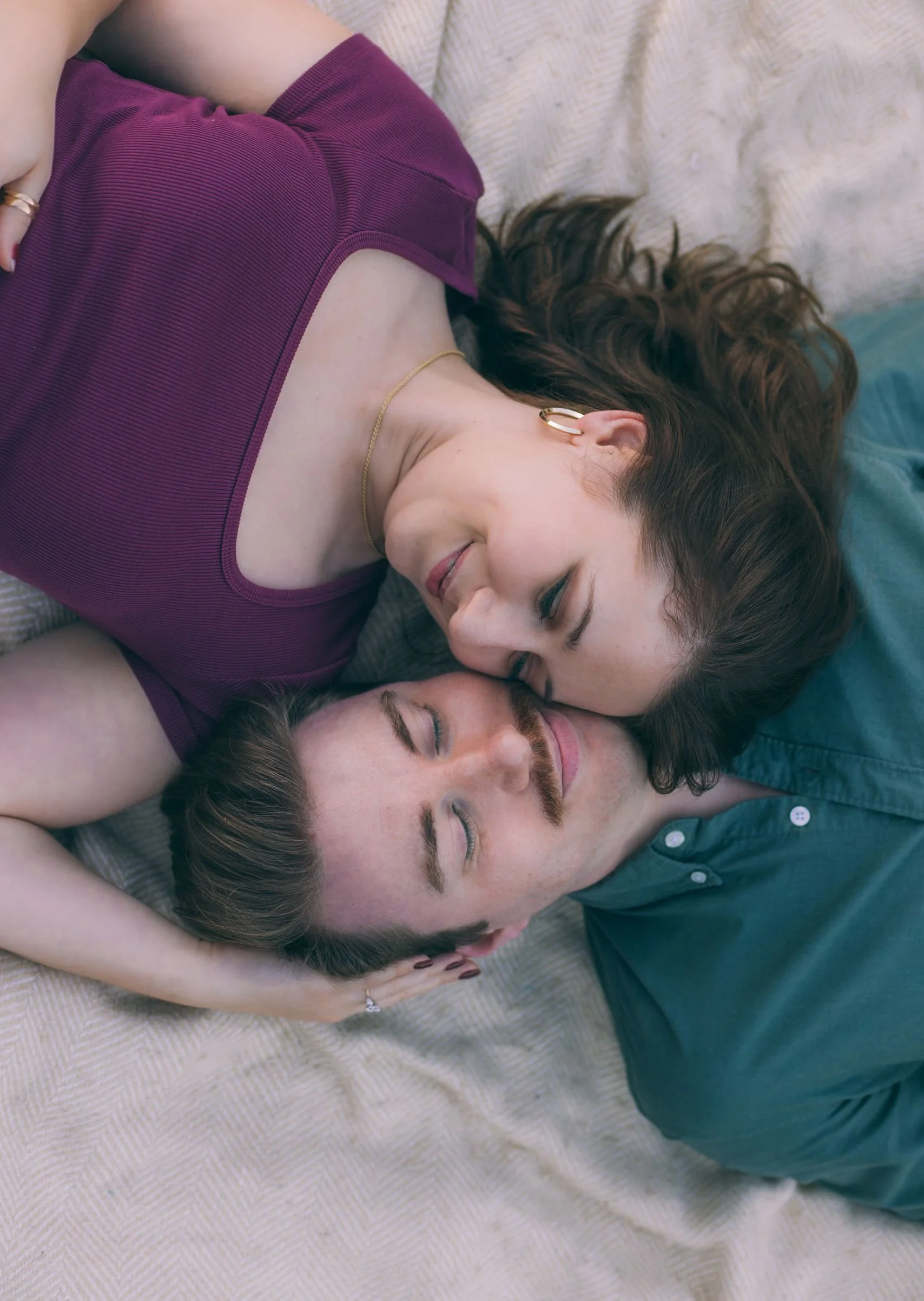 A woman and a man lying on a bed, cuddling and touching foreheads with eyes closed, smiling softly.