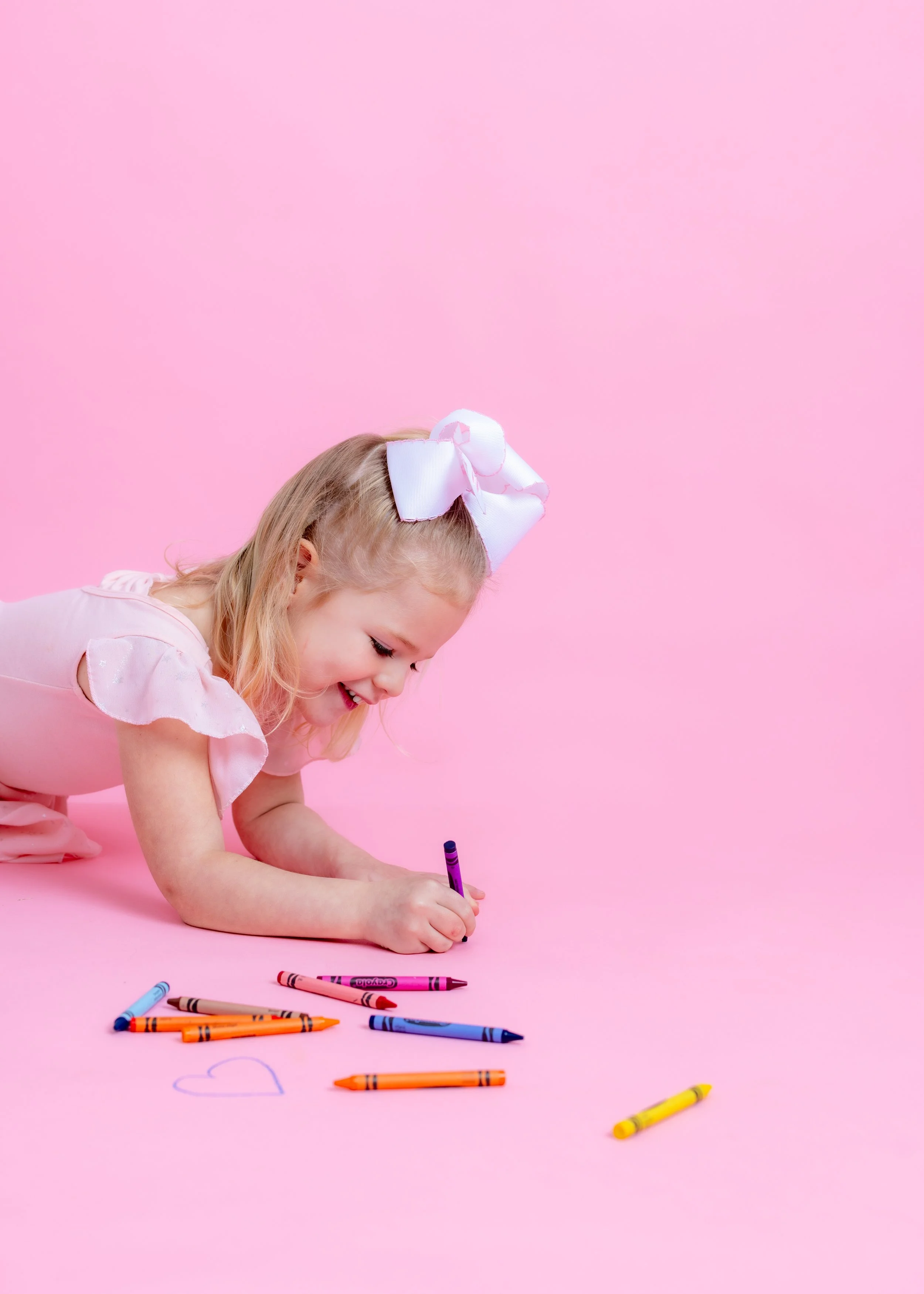A young girl with a large white bow in her hair, lying on the pink floor, drawing with crayons that are scattered around her, including a heart.