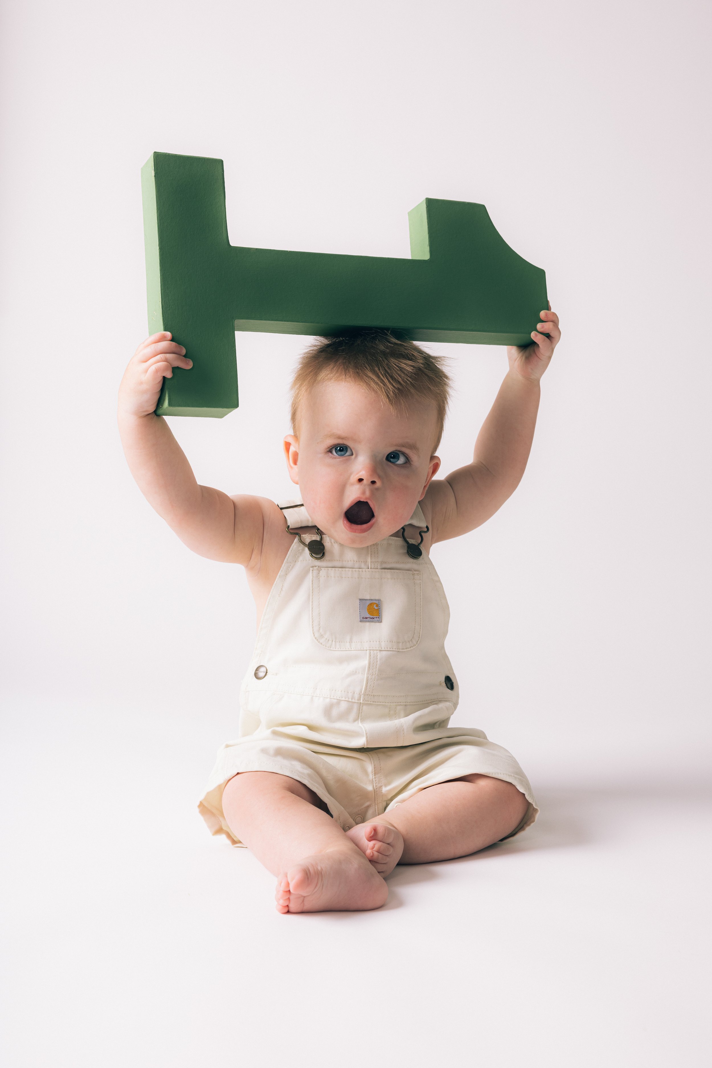 A young child with blonde hair and blue eyes, sitting on the floor in front of a plain white background, holding a large green letter H above their head with both hands, mouth open in surprise or excitement, wearing beige overalls.