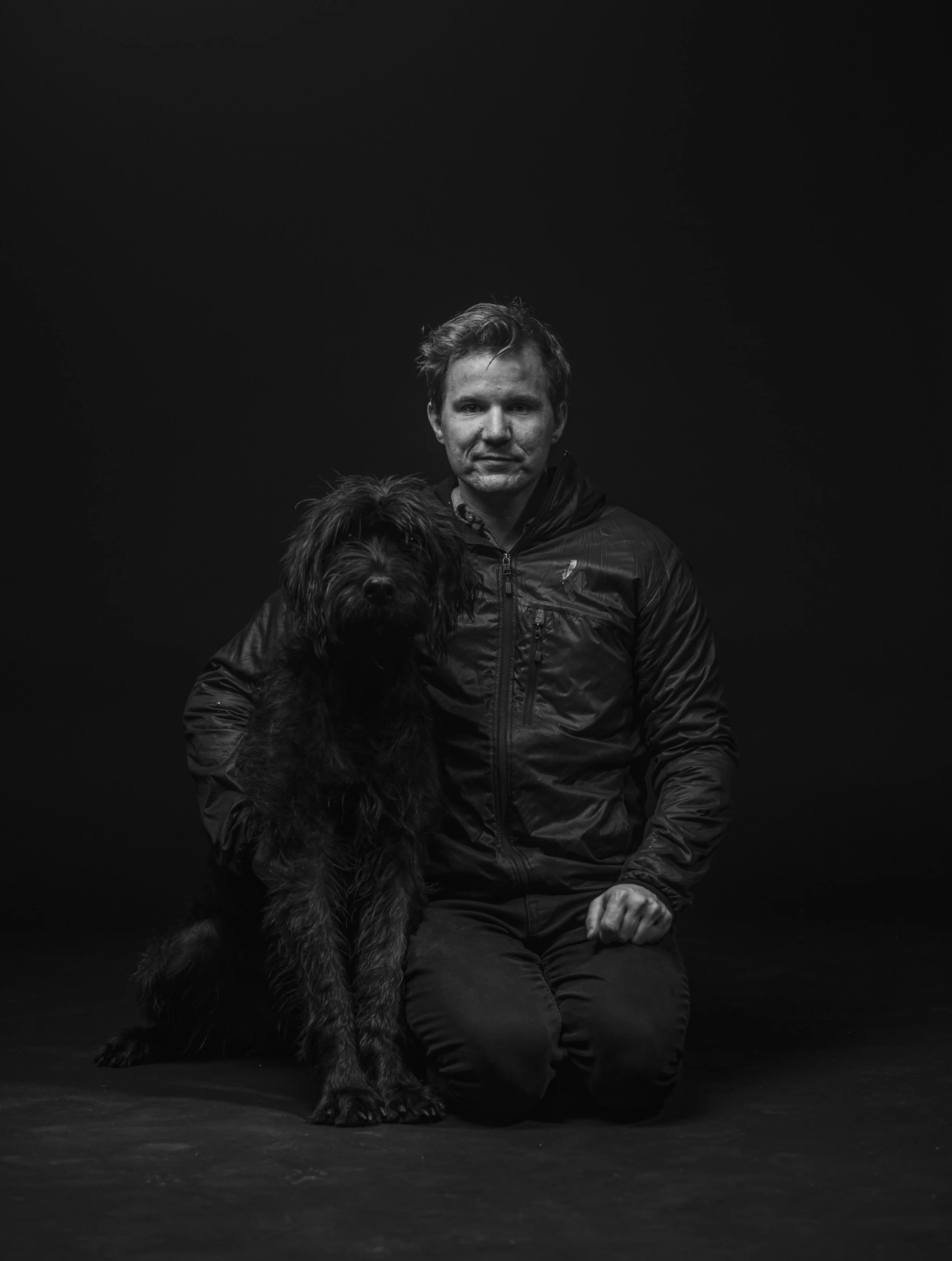 Black and white photo of a man kneeling with a large dog against a dark background.