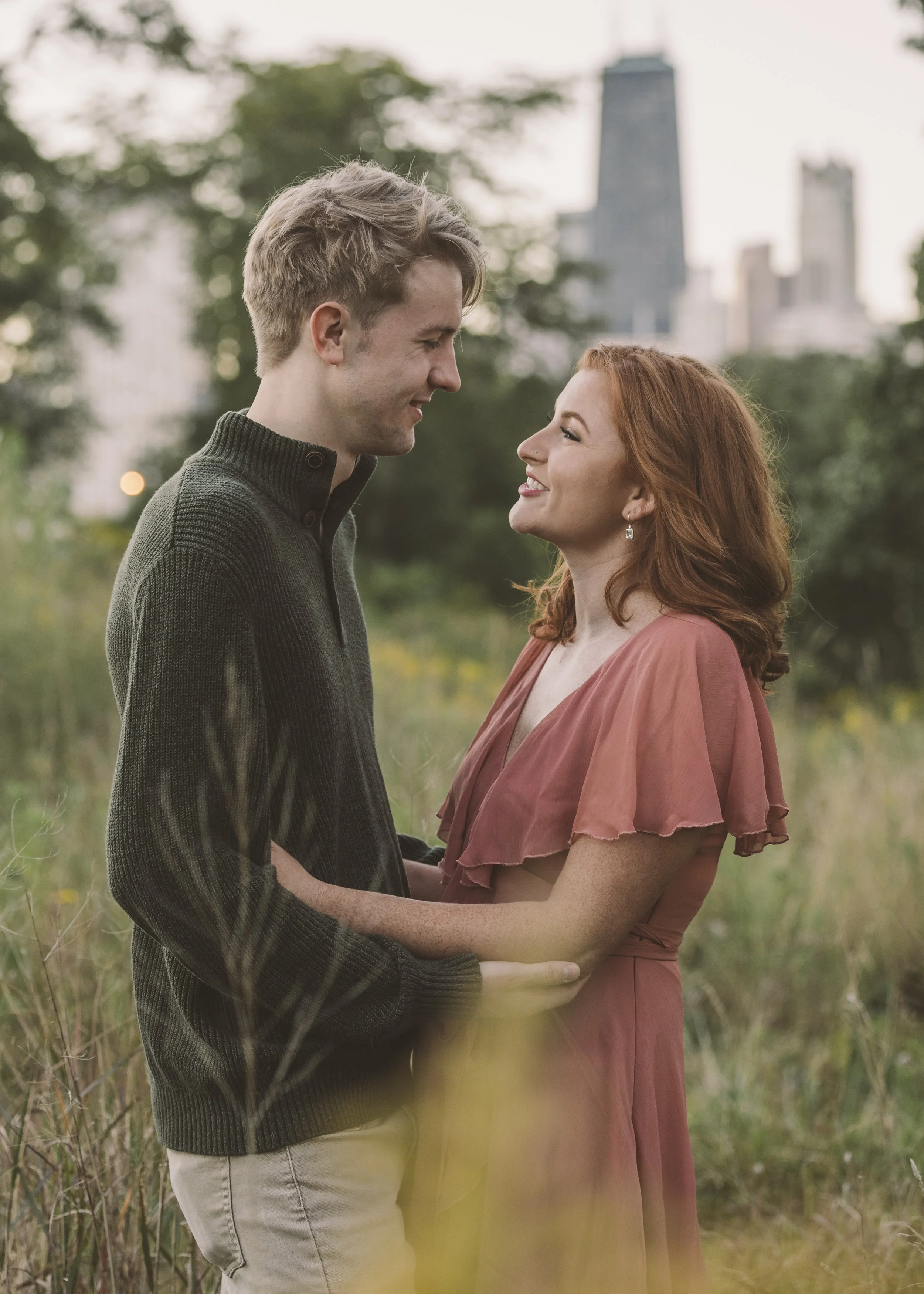 A young couple standing in a park, smiling and looking into each other's eyes, with city skyscrapers in the background.